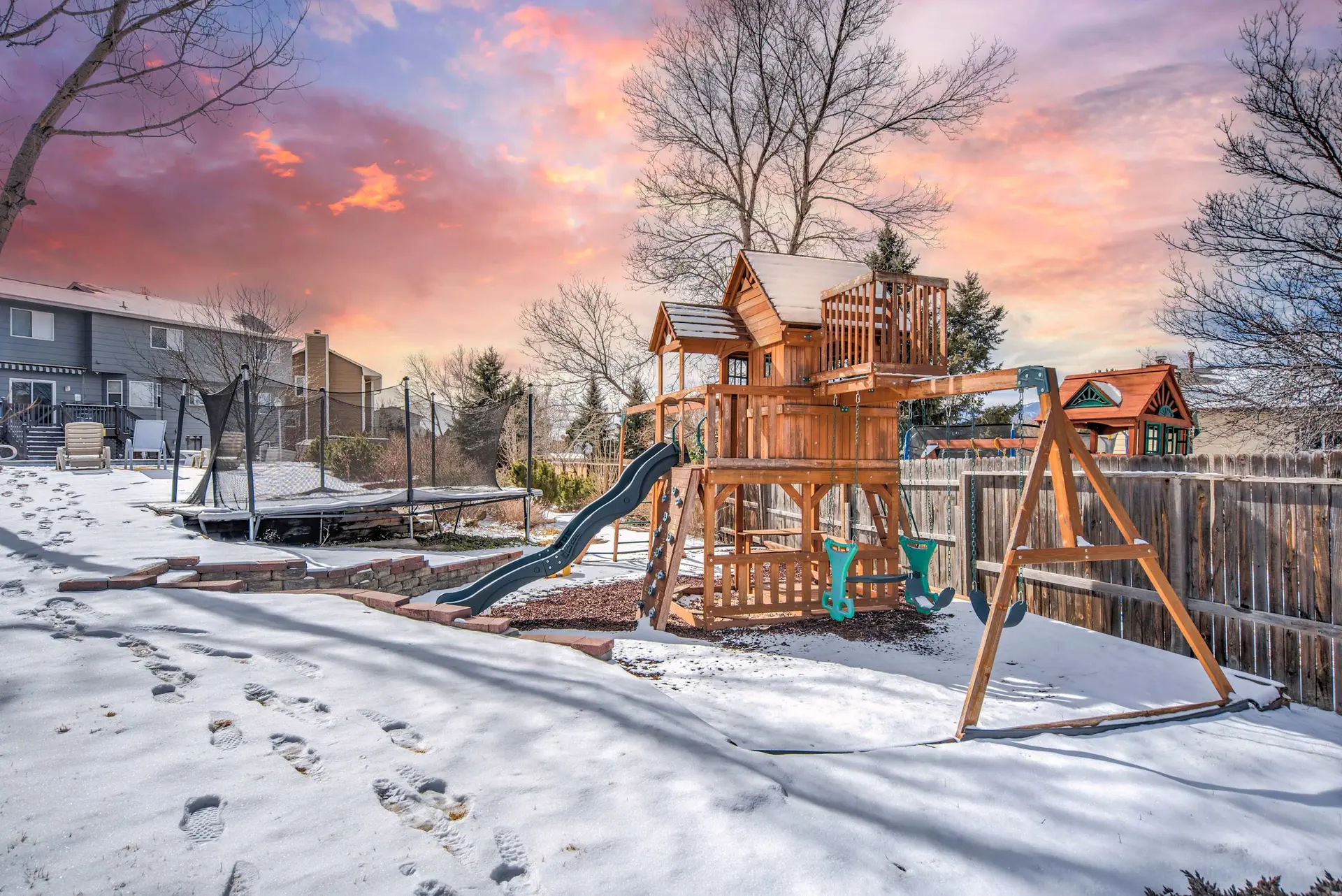 A snowy backyard with a wooden playset, trampoline, and footprints.