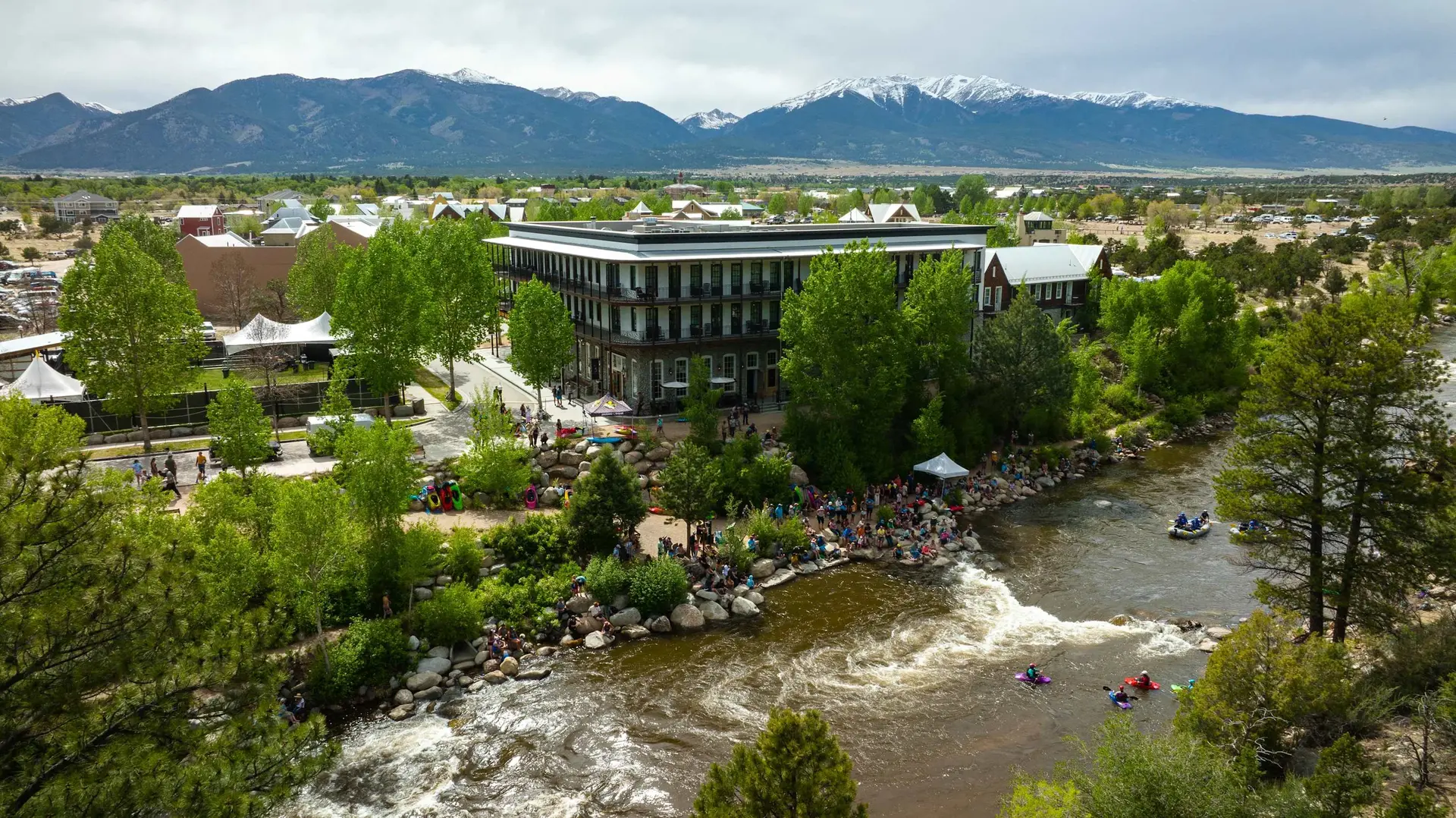 Aerial of a vibrant mountain town with a river, kayakers, spectators, and snow-capped peaks.