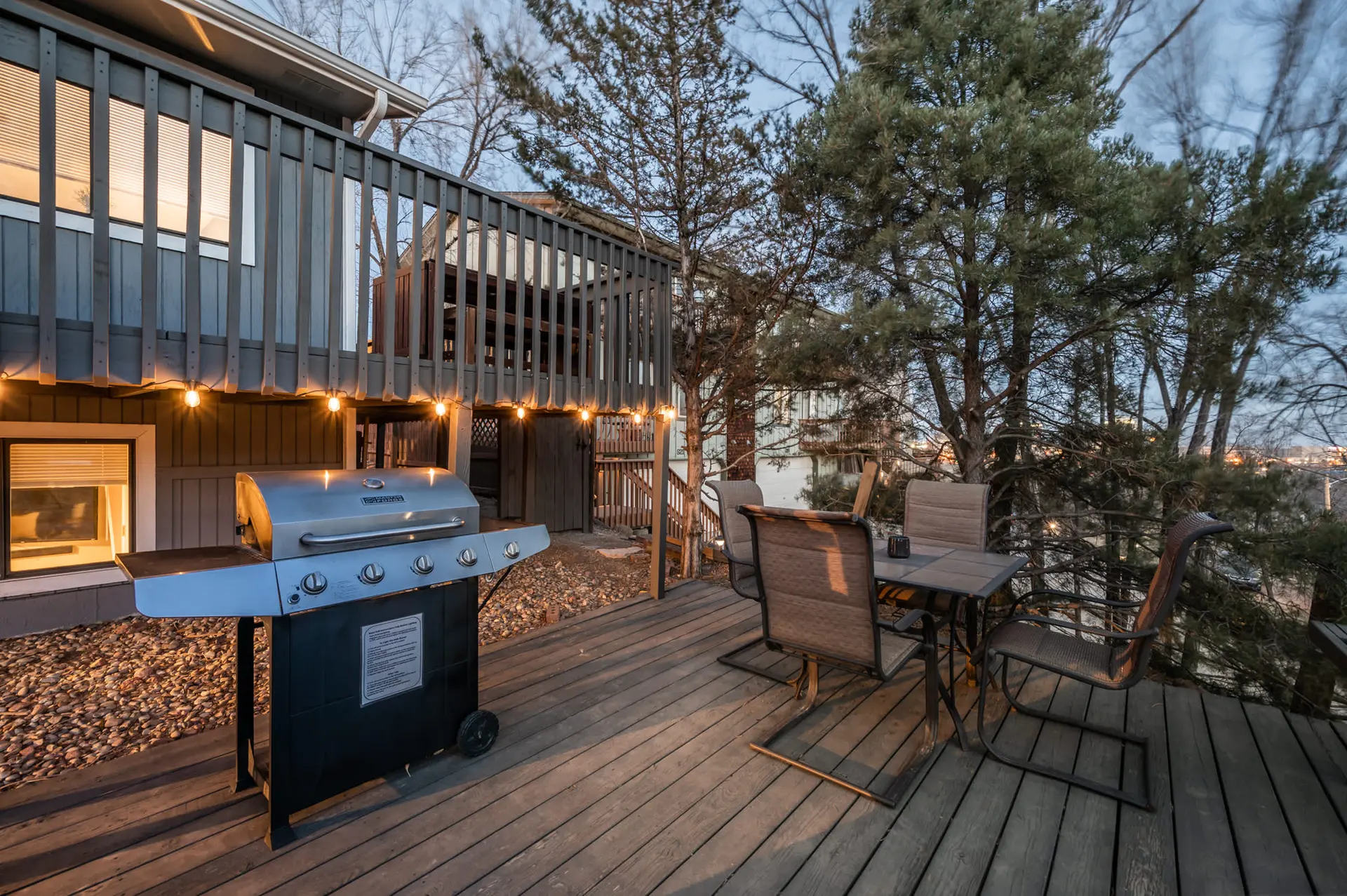 A deck with a gas grill, outdoor dining set, and string lights overlooks a wooded landscape.