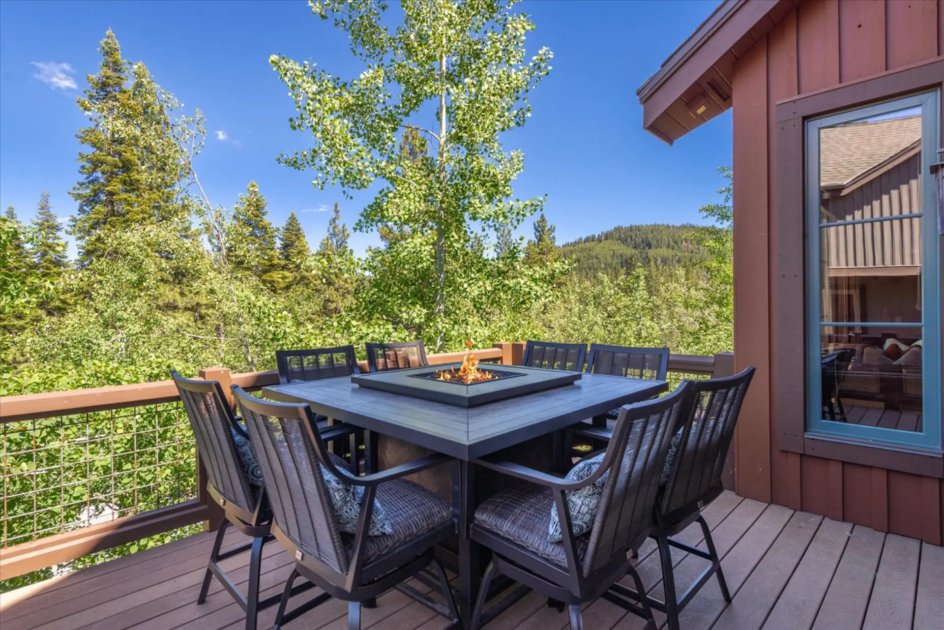 Outdoor dining set with fire pit on wooden deck surrounded by trees and mountains.