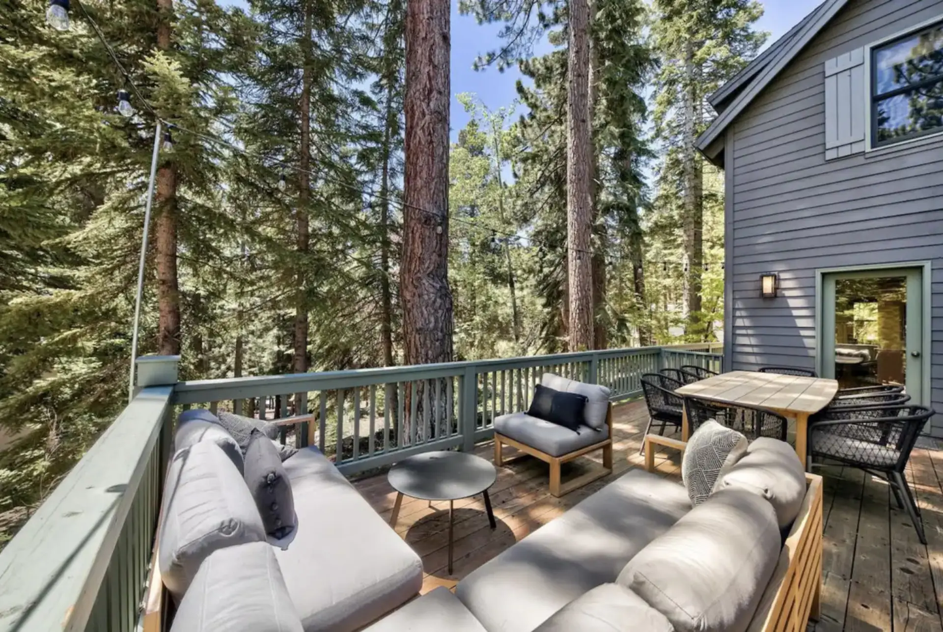 A cozy wooden deck surrounded by tall trees, featuring a sectional couch and dining set.