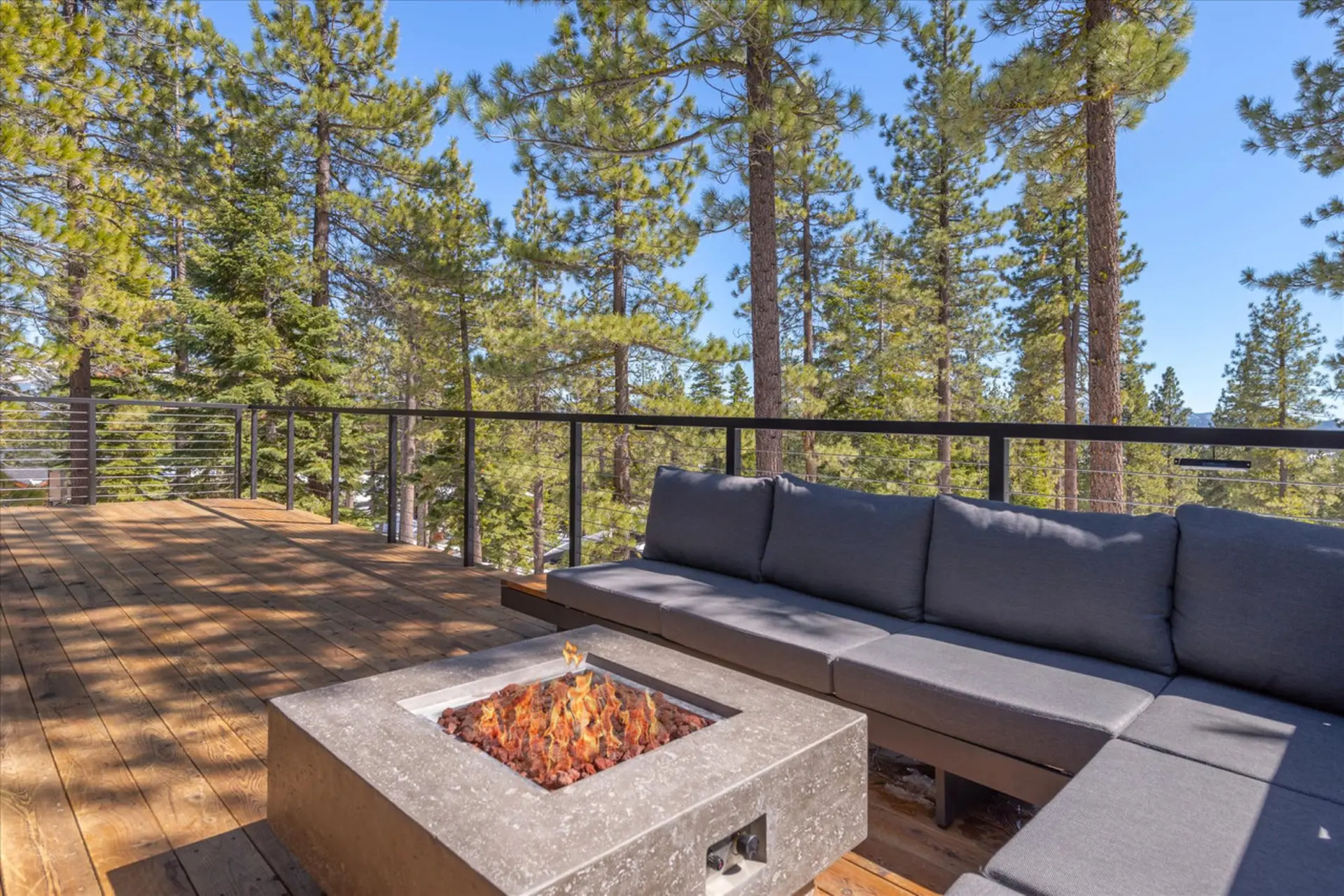 A deck with a fire pit and sectional sofa surrounded by pine trees.