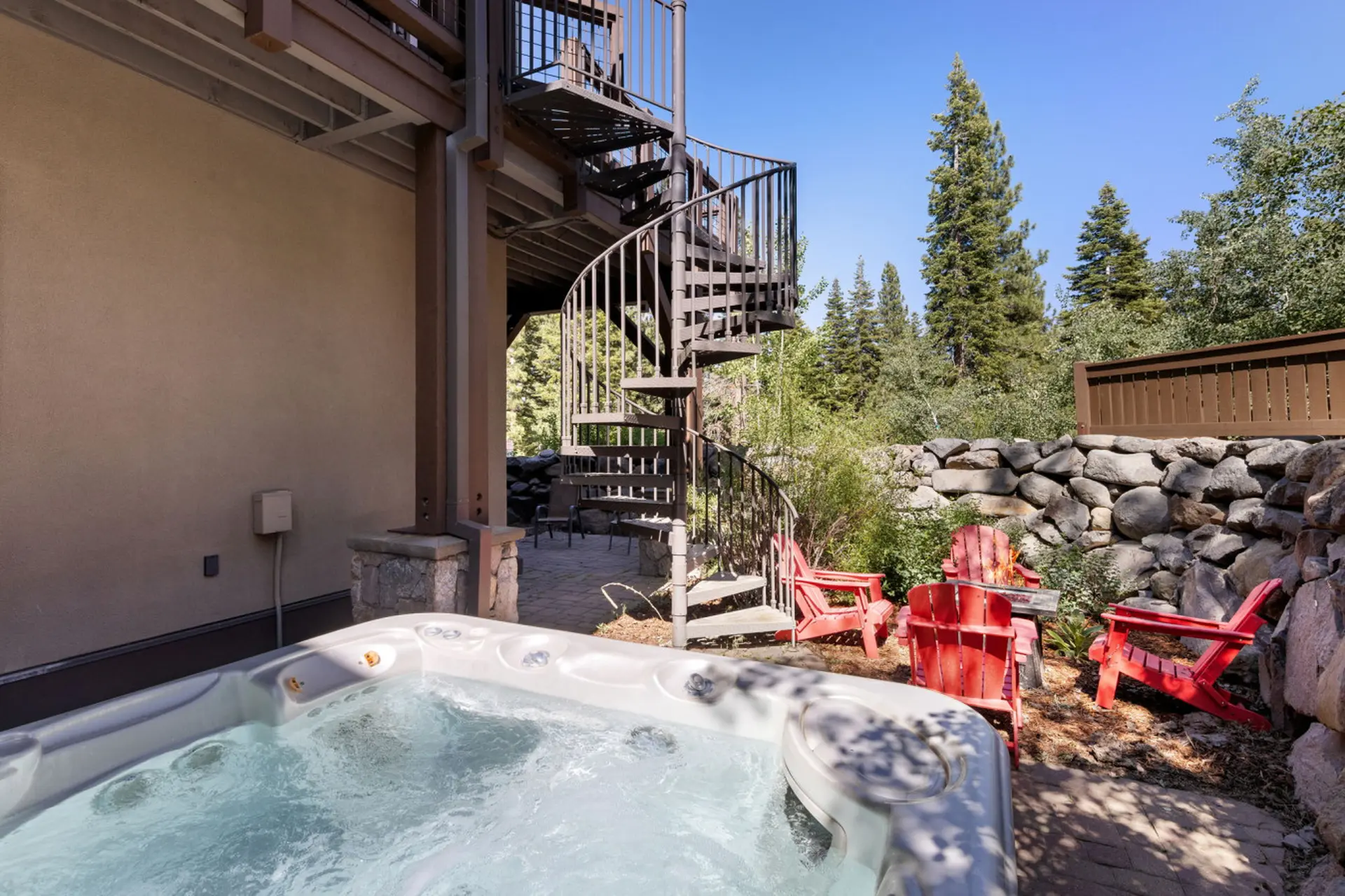 An outdoor hot tub and red Adirondack chairs sit on a stone patio.