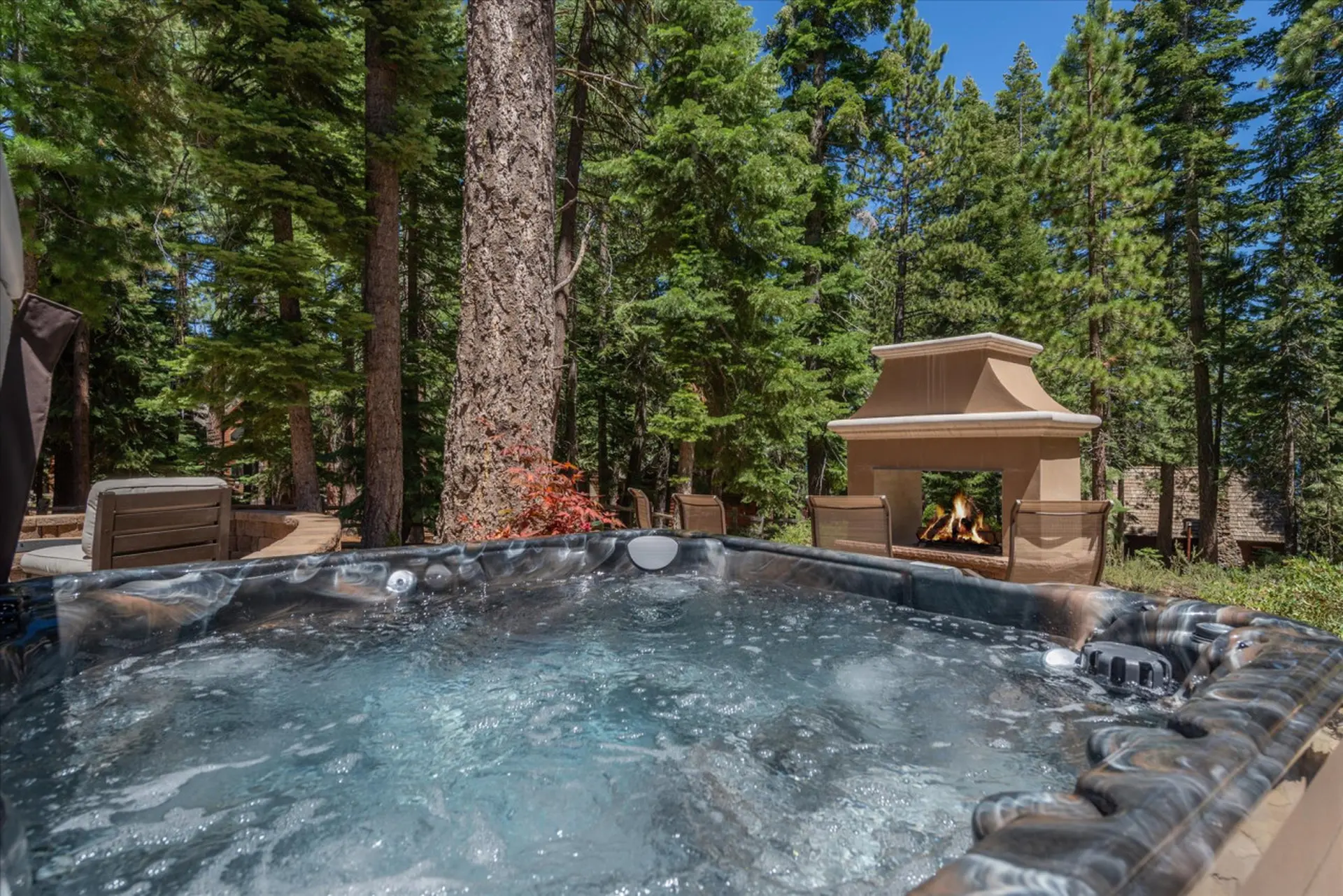 A hot tub sits in front of an outdoor fireplace, surrounded by tall trees.