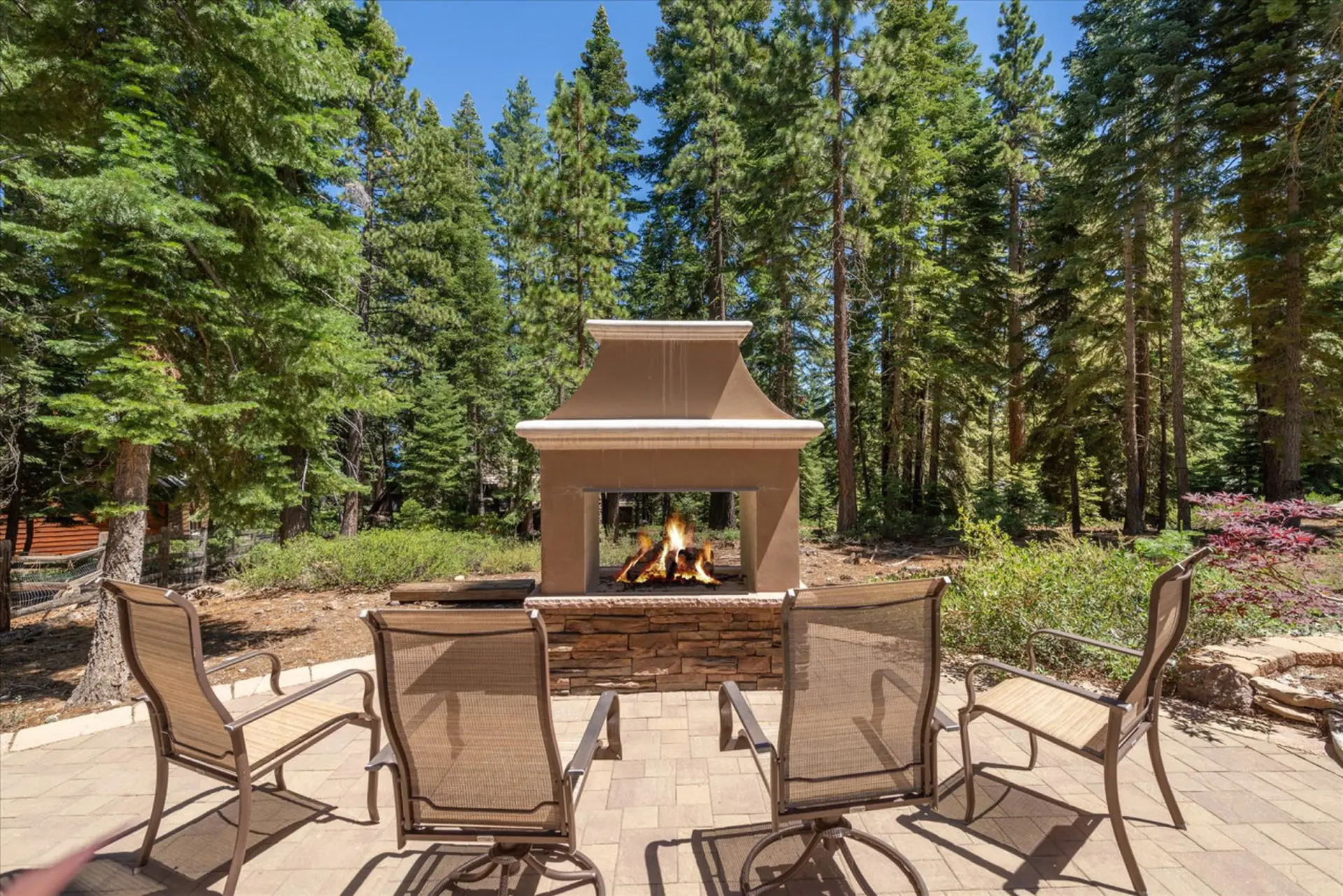 Outdoor fireplace with seating on a stone patio surrounded by pine trees.
