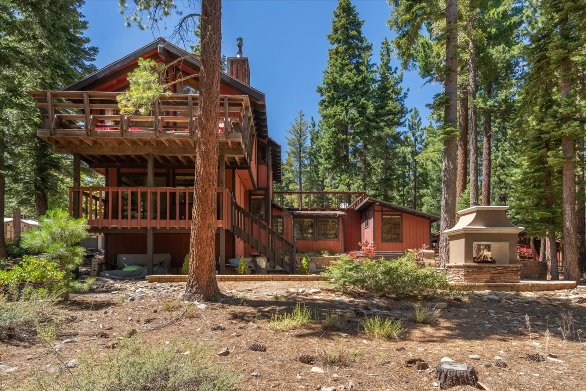 Rustic red cabin nestled among tall pine trees with a wooden deck and outdoor fireplace.