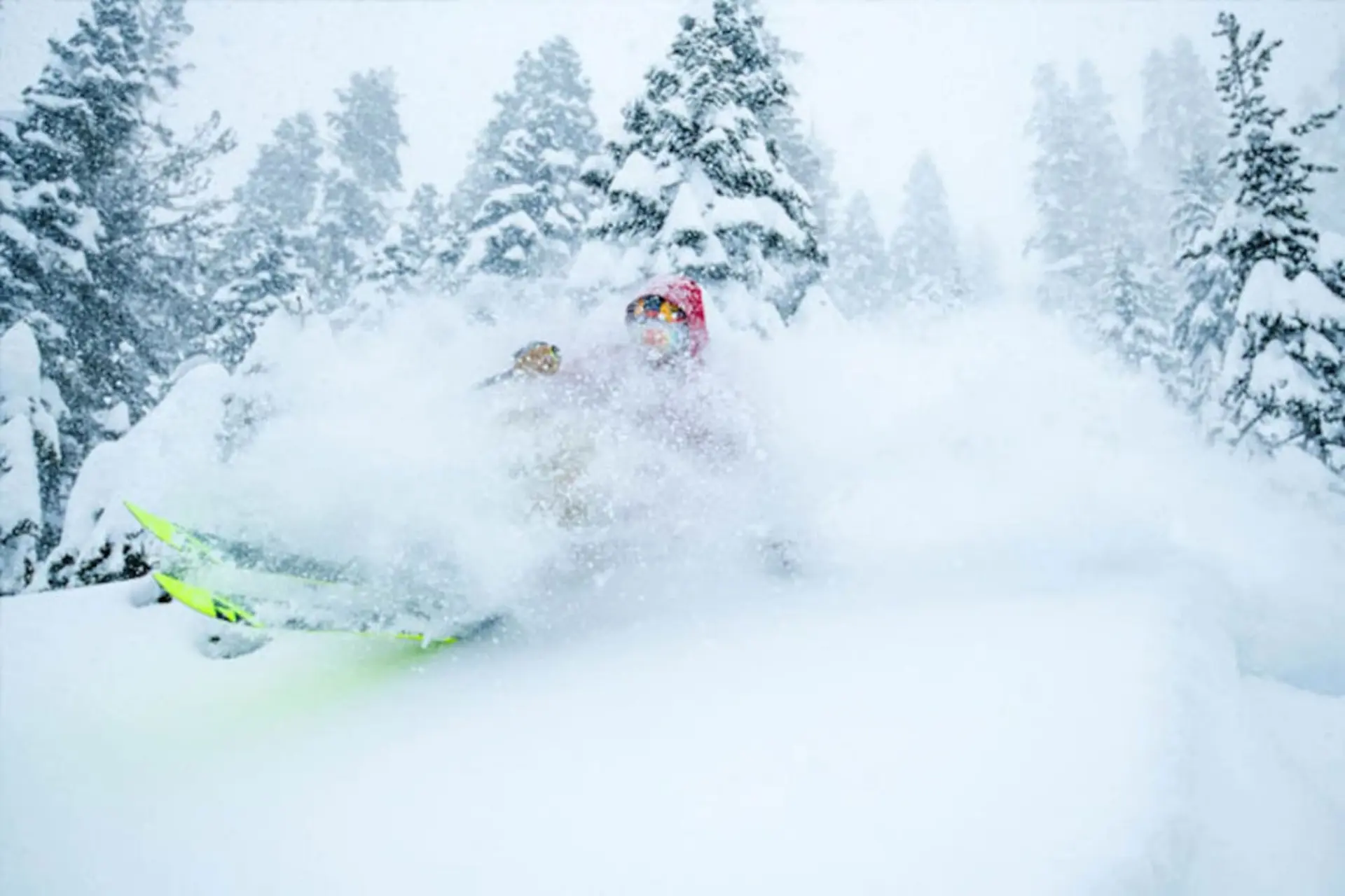 A skier in a red hat carves through deep powder snow, creating a large spray.