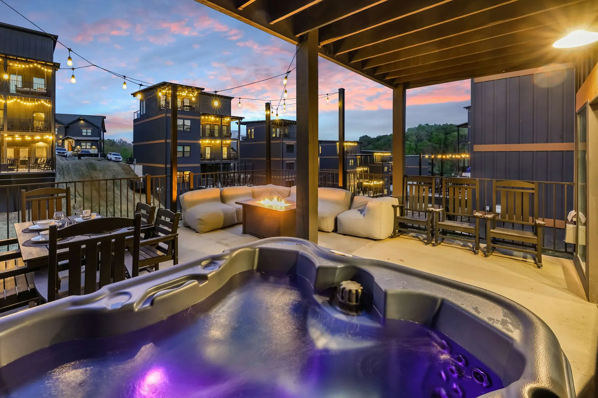 A covered balcony with a hot tub, fire pit, seating, and a dining table overlooks modern buildings a