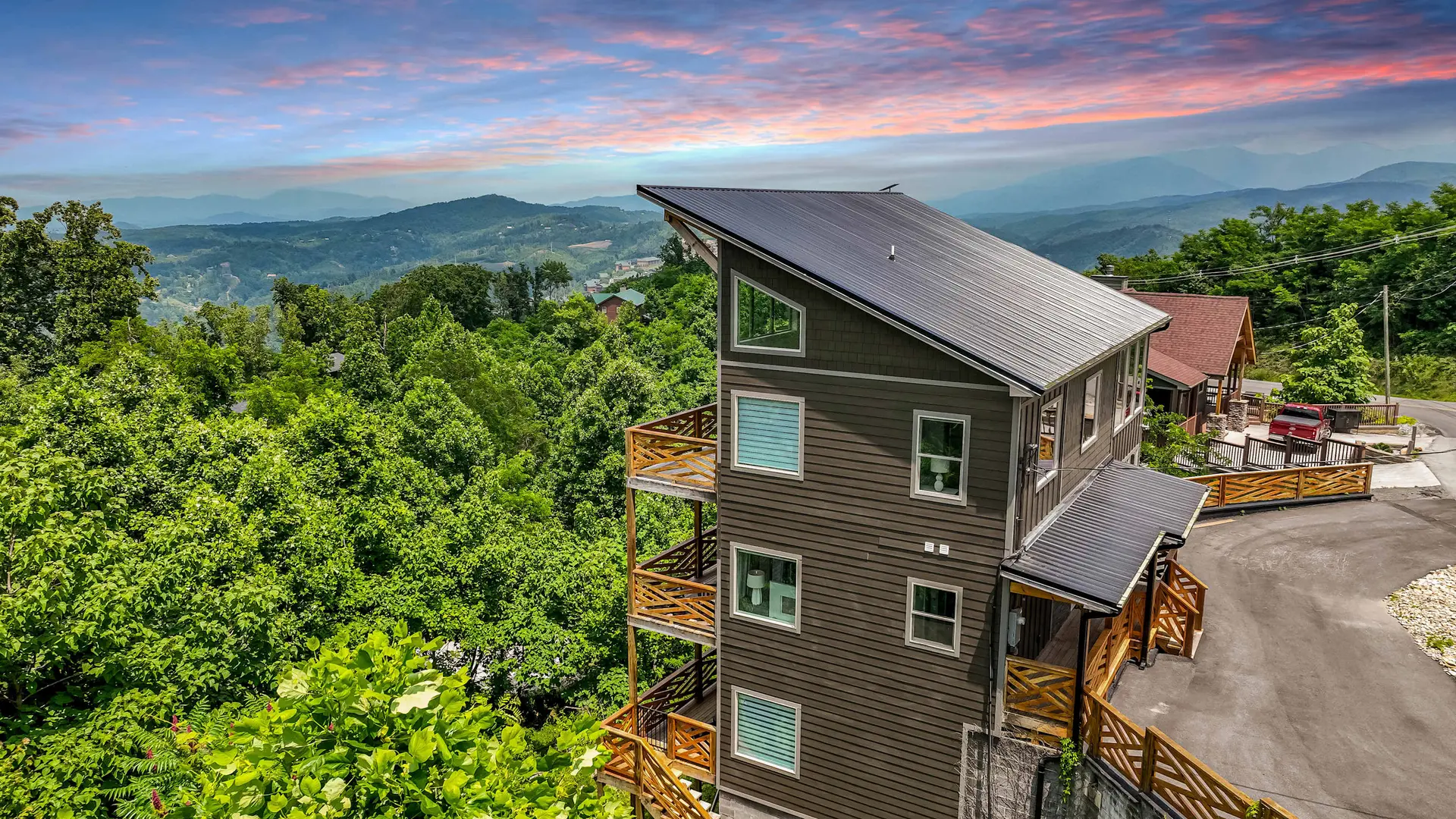 A modern brown house sits on a wooded mountainside under a colorful sunset.