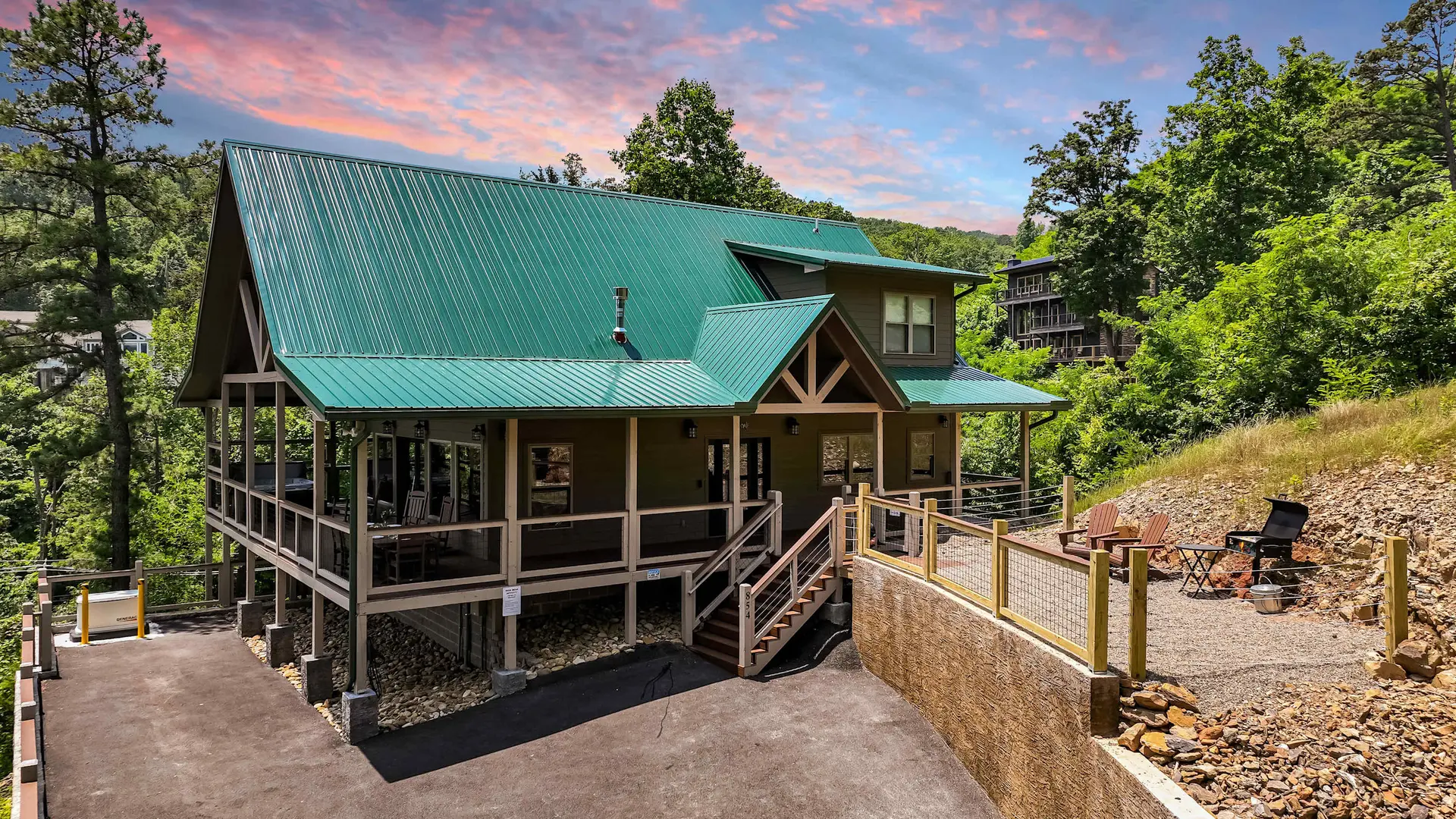 A wooden cabin with a green metal roof sits on a wooded hill. There is a deck with seating and a gri