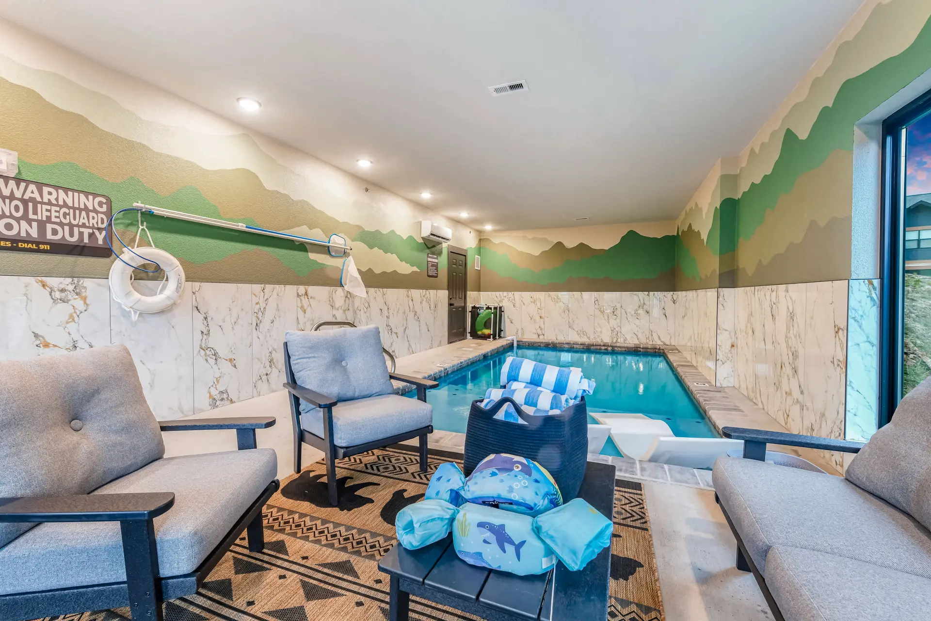 Indoor swimming pool area with chairs, a table with towels and floaties, and a mountain mural.