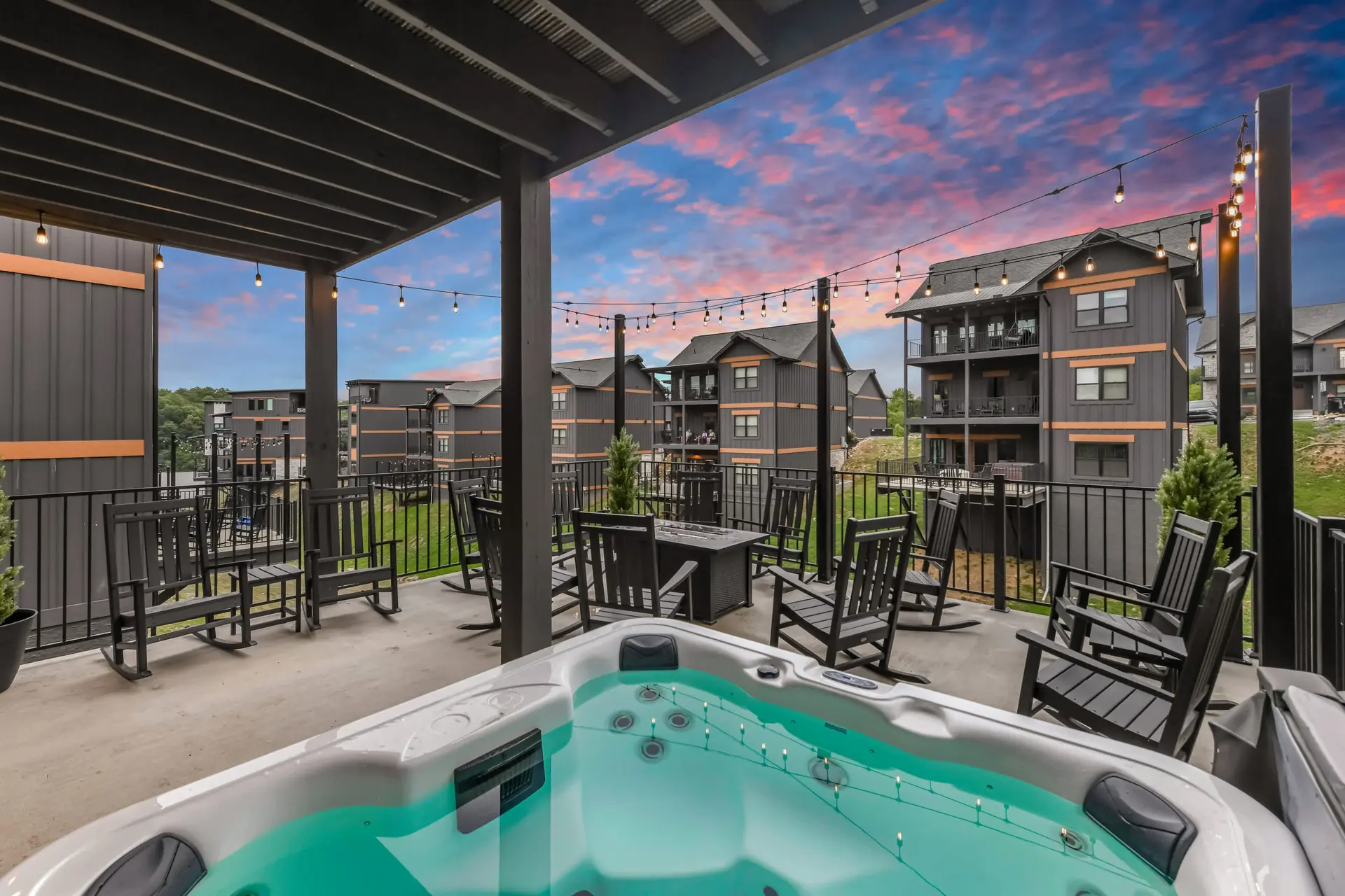 Hot tub on a patio with rocking chairs and string lights, overlooking buildings at sunset.