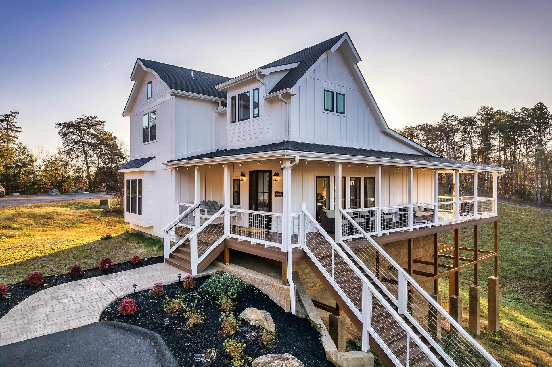 Modern white farmhouse with porch and stairs, surrounded by trees and grass.