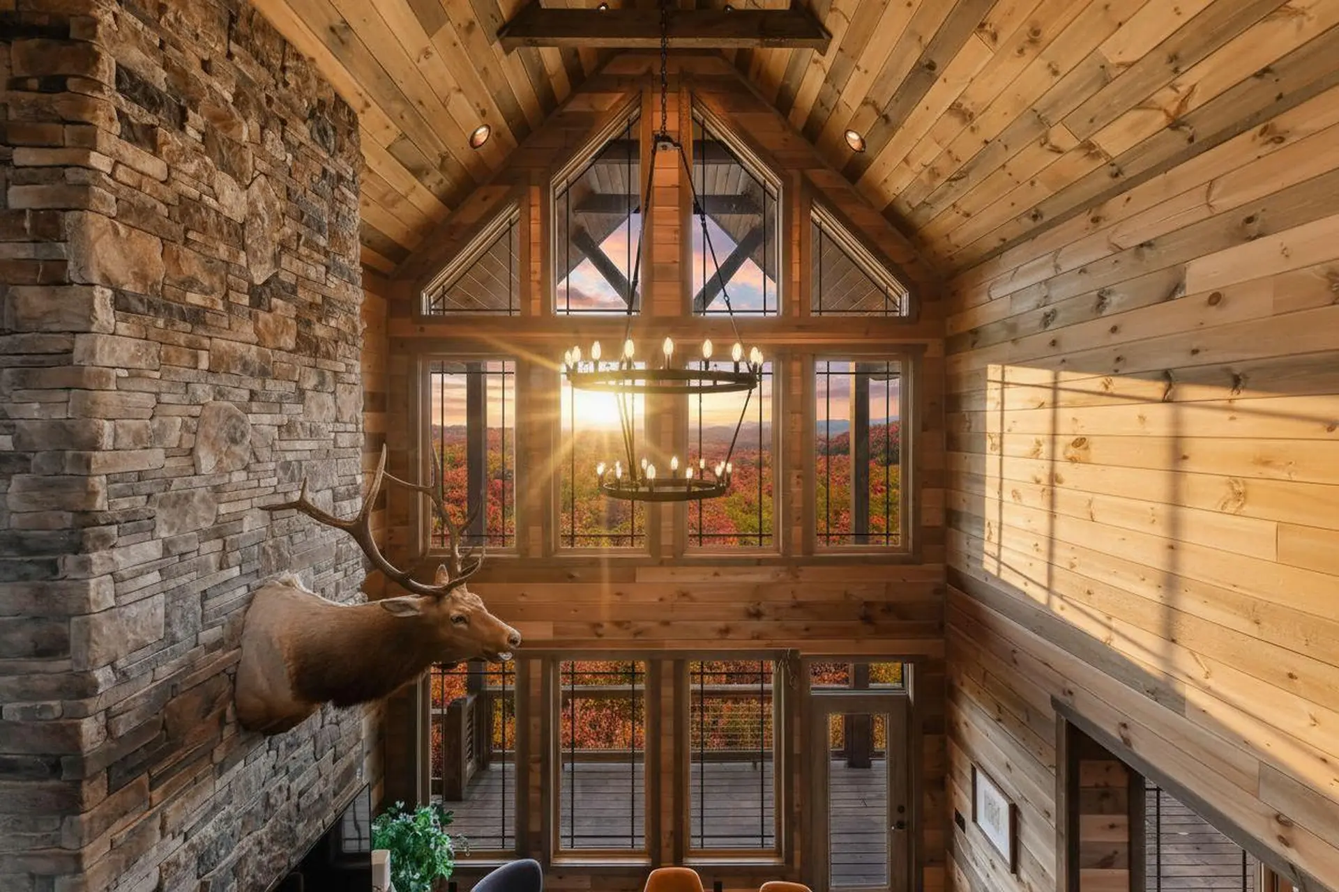 Deer head mount on stone fireplace in rustic room with large windows overlooking autumn forest.