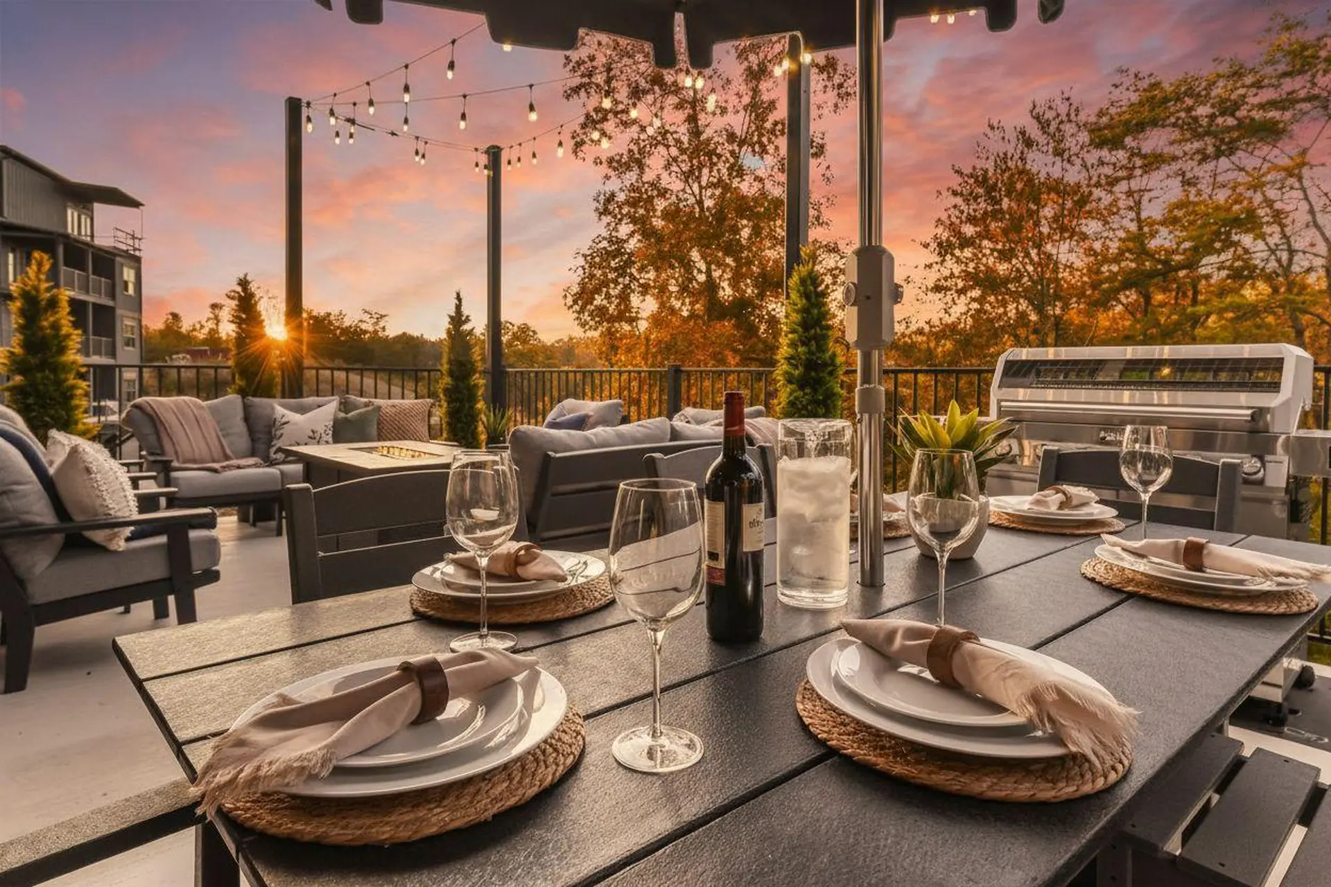 An outdoor dining set up with string lights, a grill, and a sunset view.