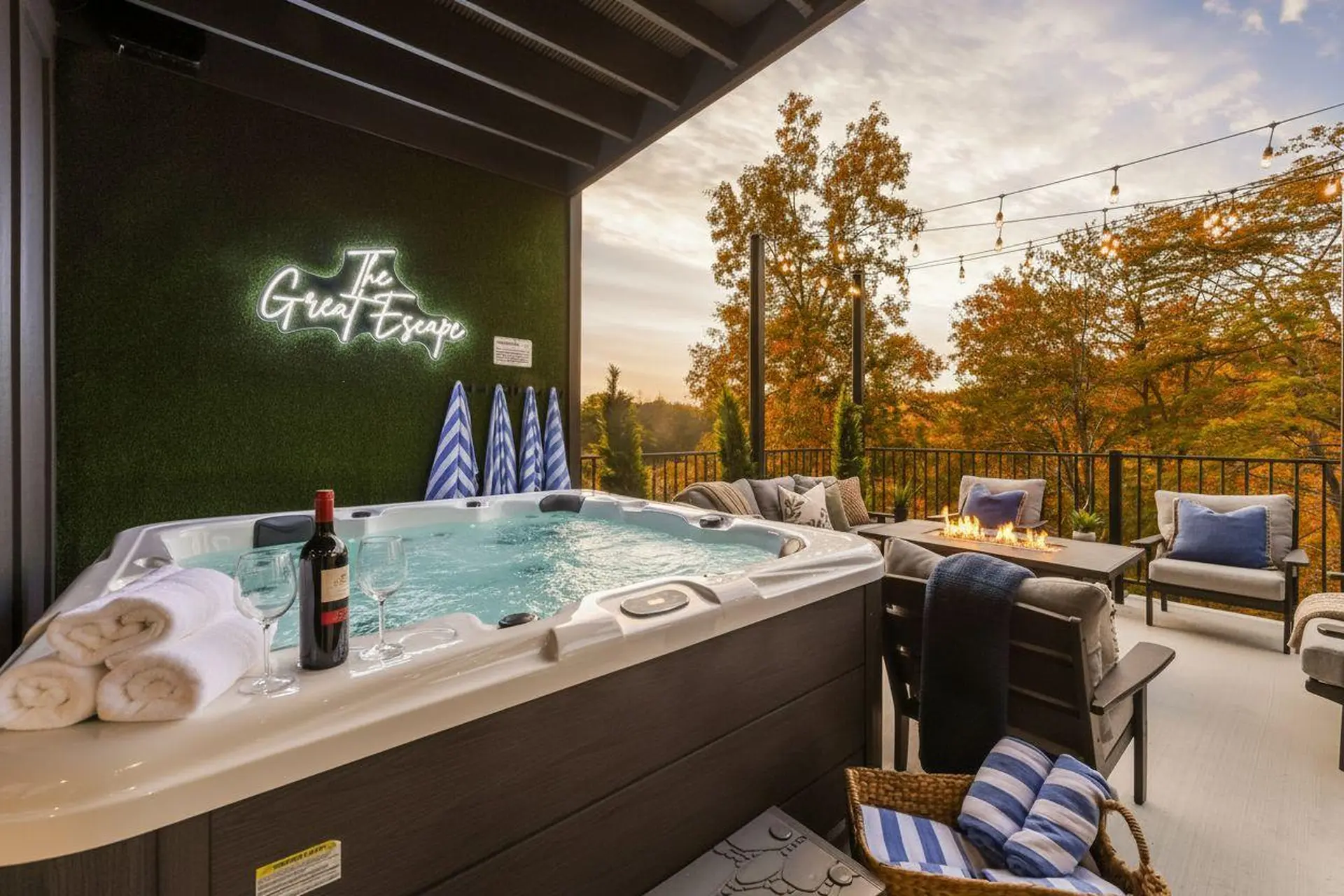 Hot tub on a deck with a fire pit, chairs, and string lights overlooking autumn foliage.