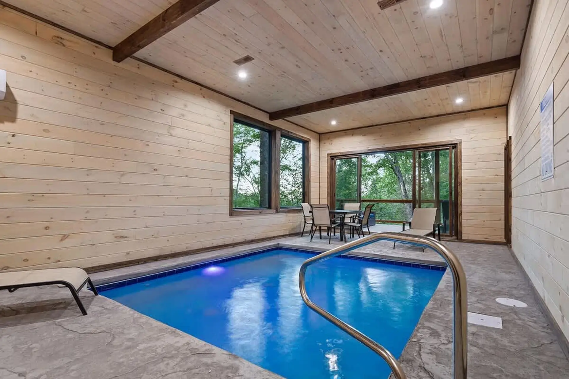Indoor pool with lounge chairs and dining area overlooking a wooded backyard.
