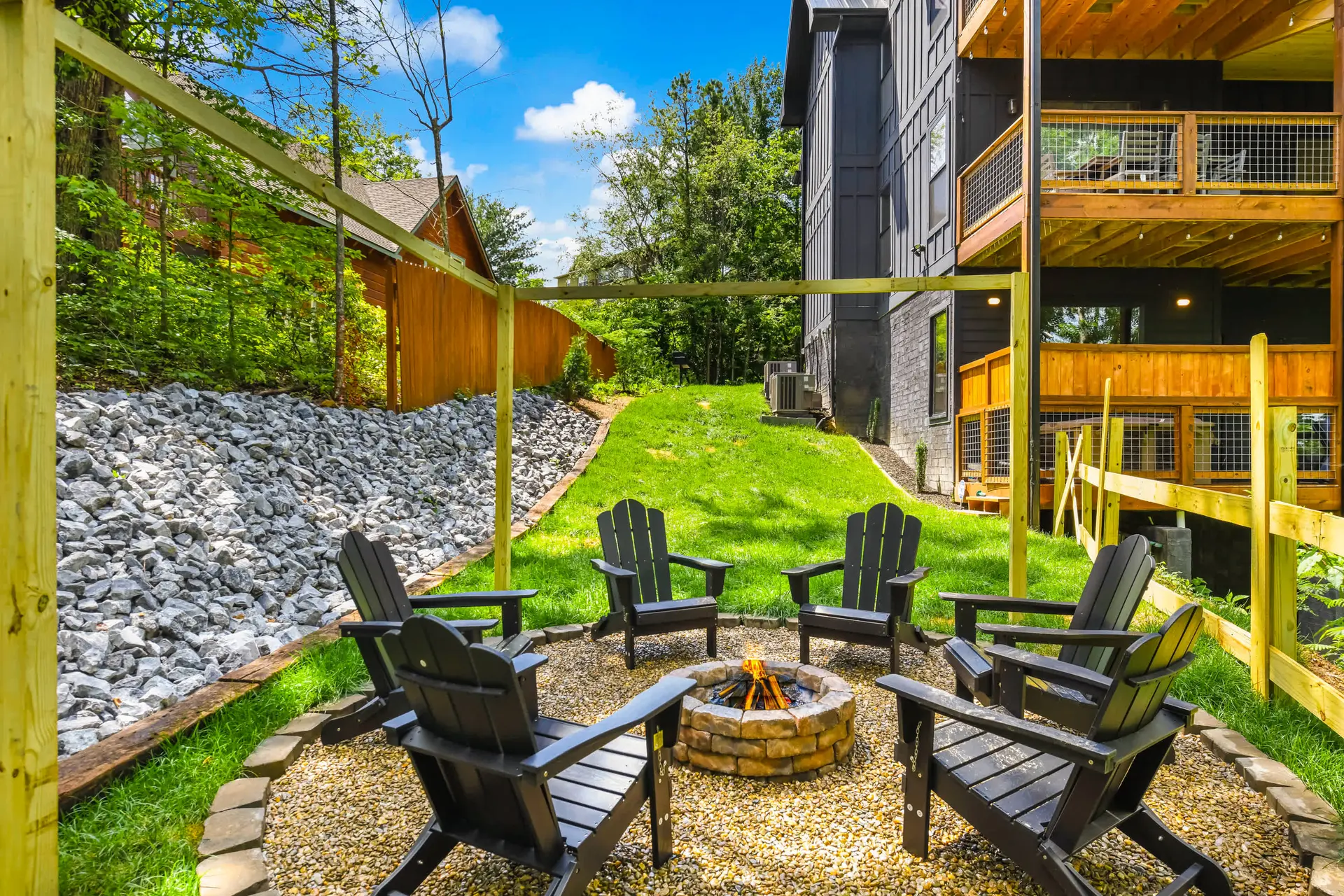 A fire pit with Adirondack chairs in a grassy backyard, surrounded by trees and a modern building.