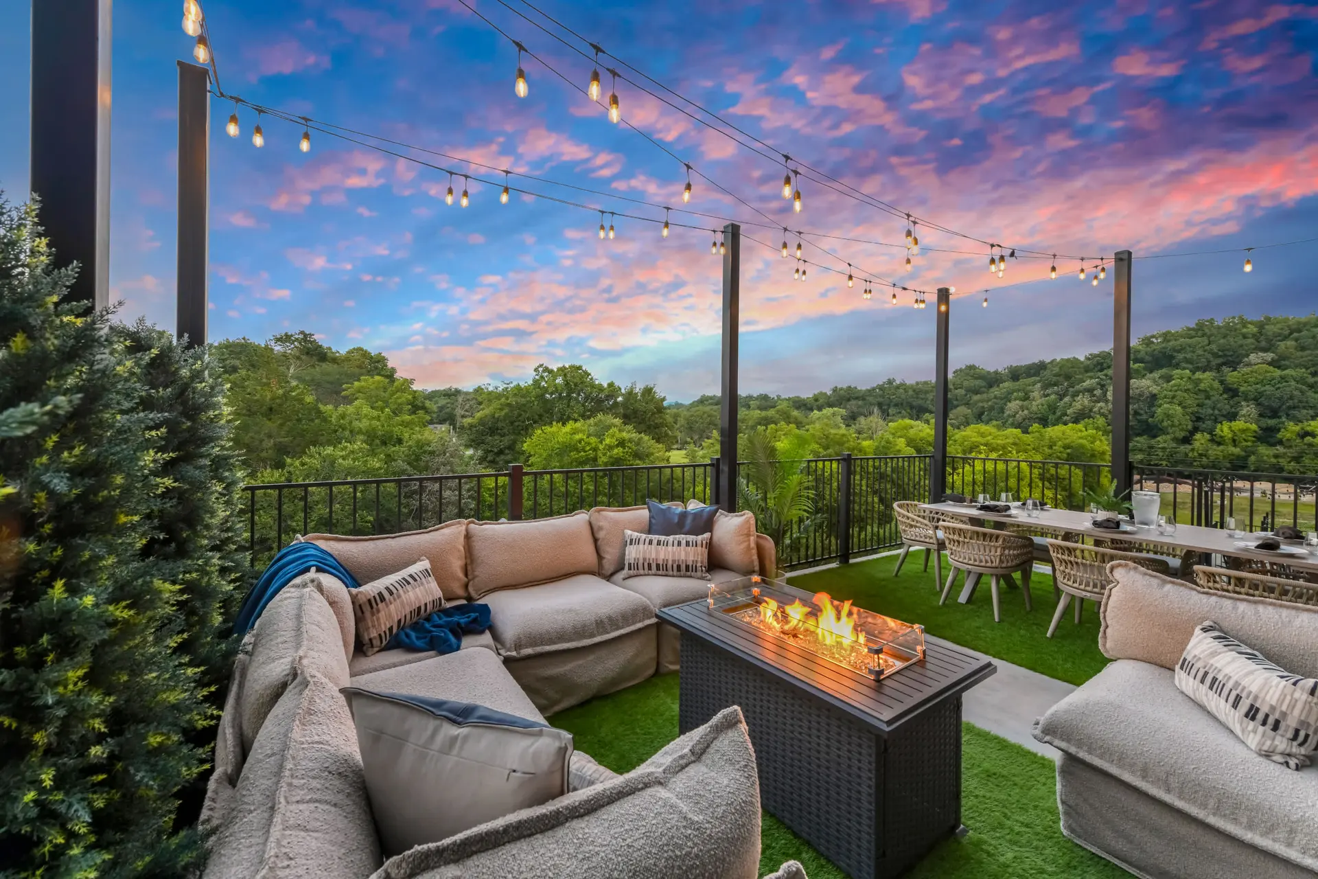 An outdoor patio at dusk with a sectional sofa, fire pit, and dining table, set against a colorful s