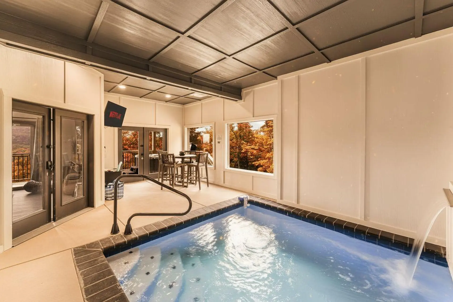 Indoor hot tub with a view of autumn trees, a TV, and a bar area.
