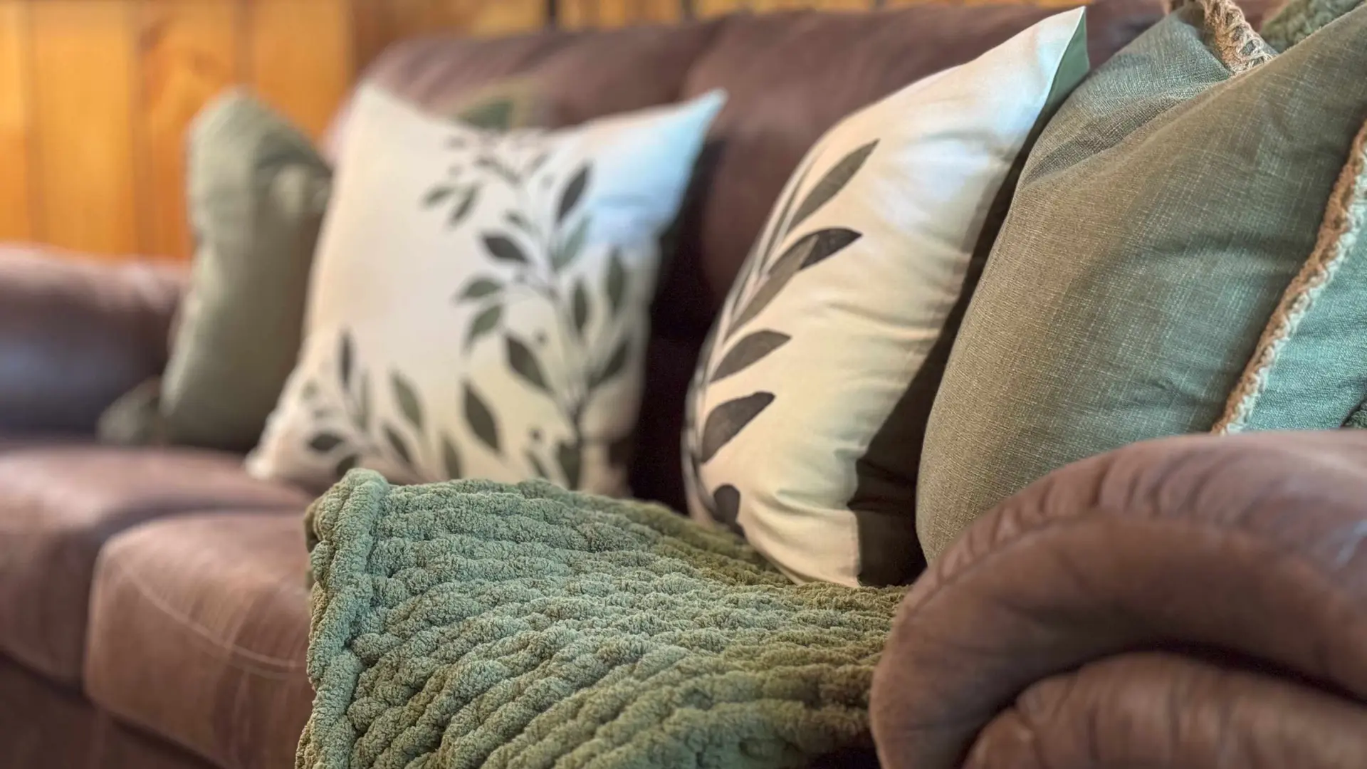 A cozy brown sofa with green pillows and a chunky knit blanket.