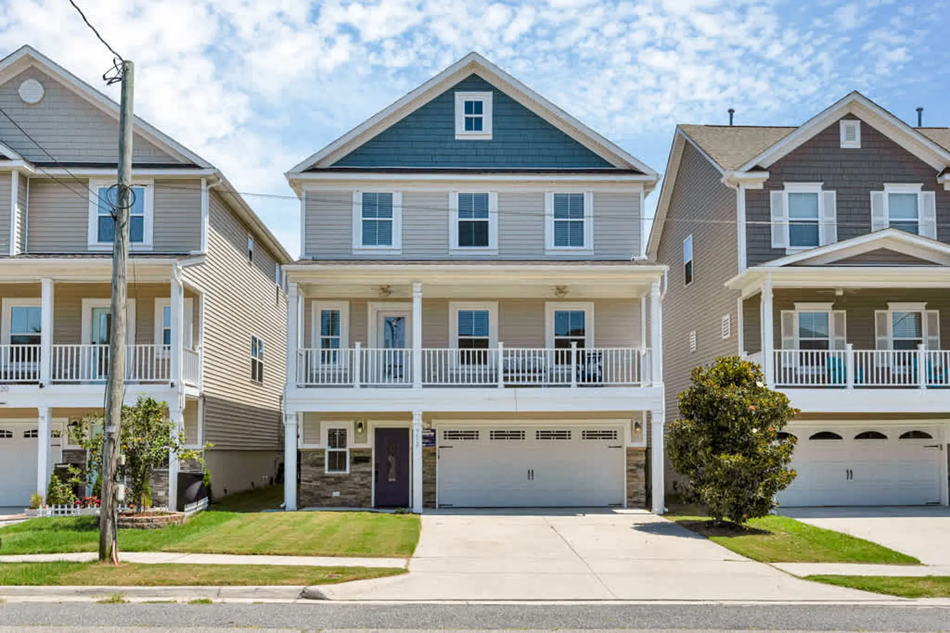 A multi-story beige house with a blue gable, white porch, and two-car garage on a sunny day.