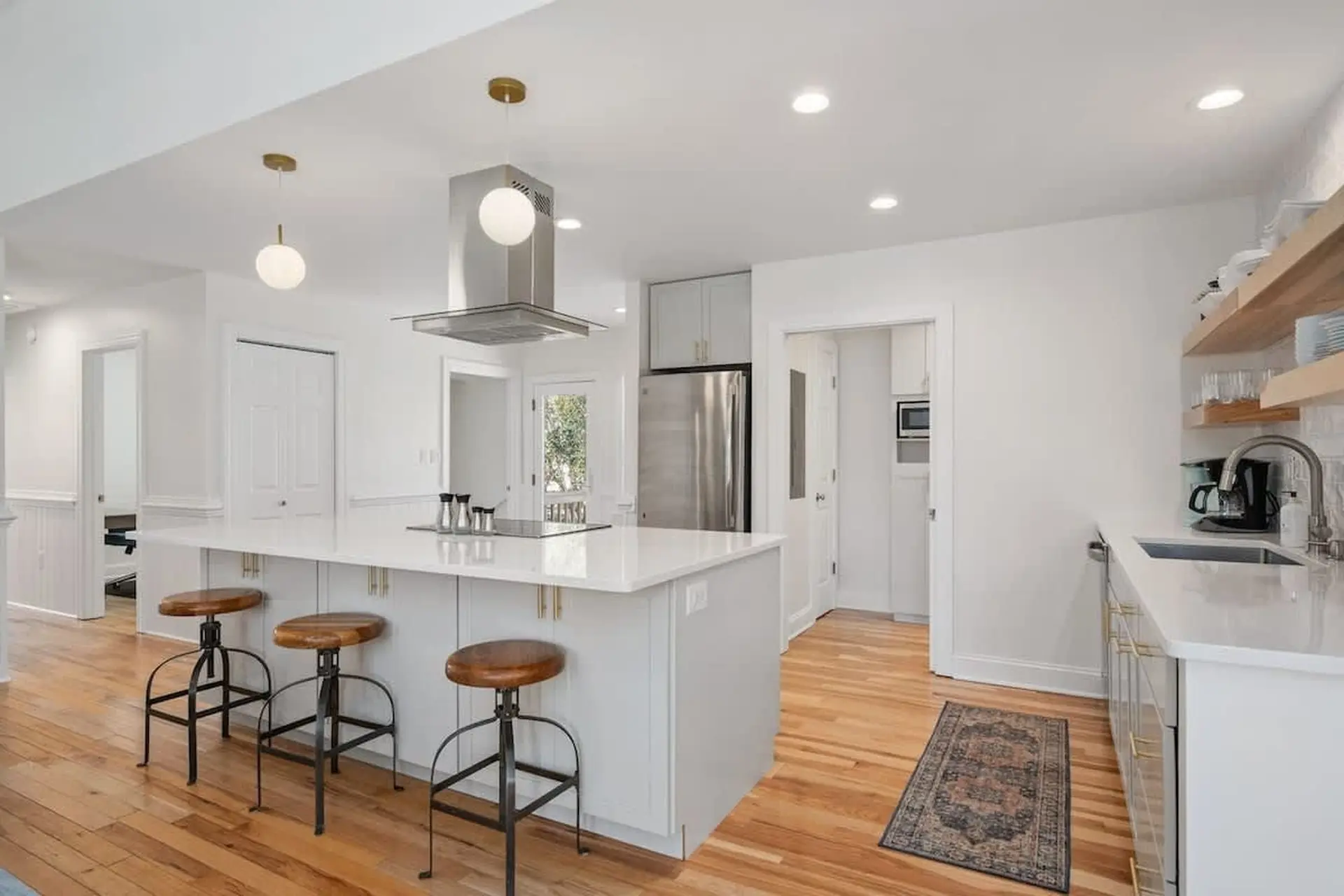 Modern white kitchen with island, stools, stainless steel appliances, and wooden floors.