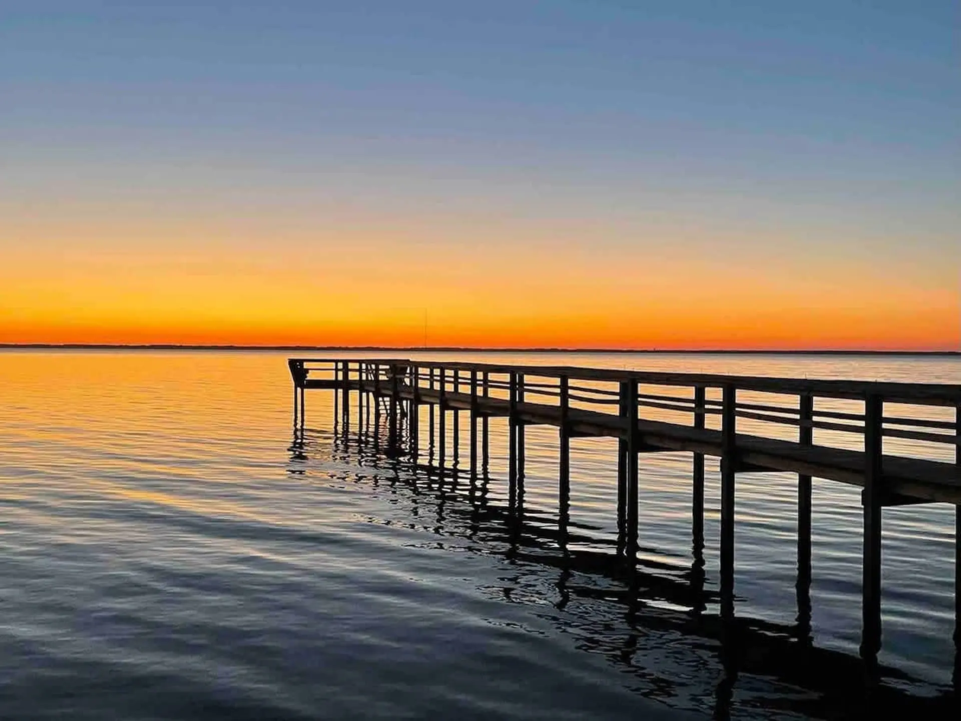 A wooden pier extends into calm water during a vibrant orange and blue sunset.