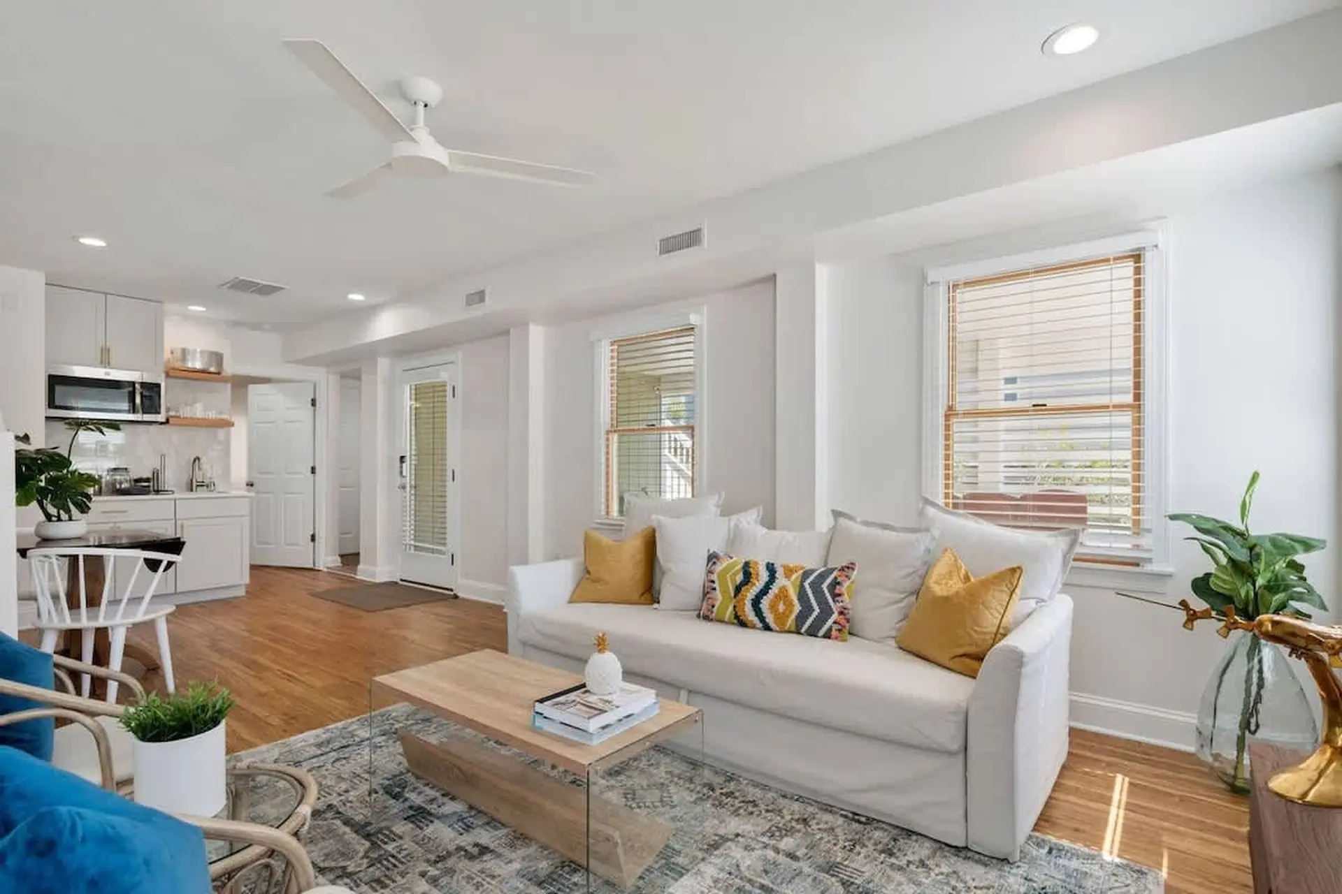 A bright, airy living room and kitchen with a white sofa, wooden furniture, and a ceiling fan.