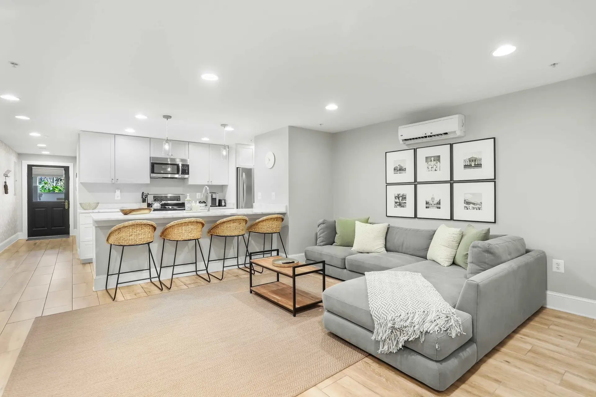 Modern living room and kitchen with grey sectional sofa, wicker bar stools, and a black door.