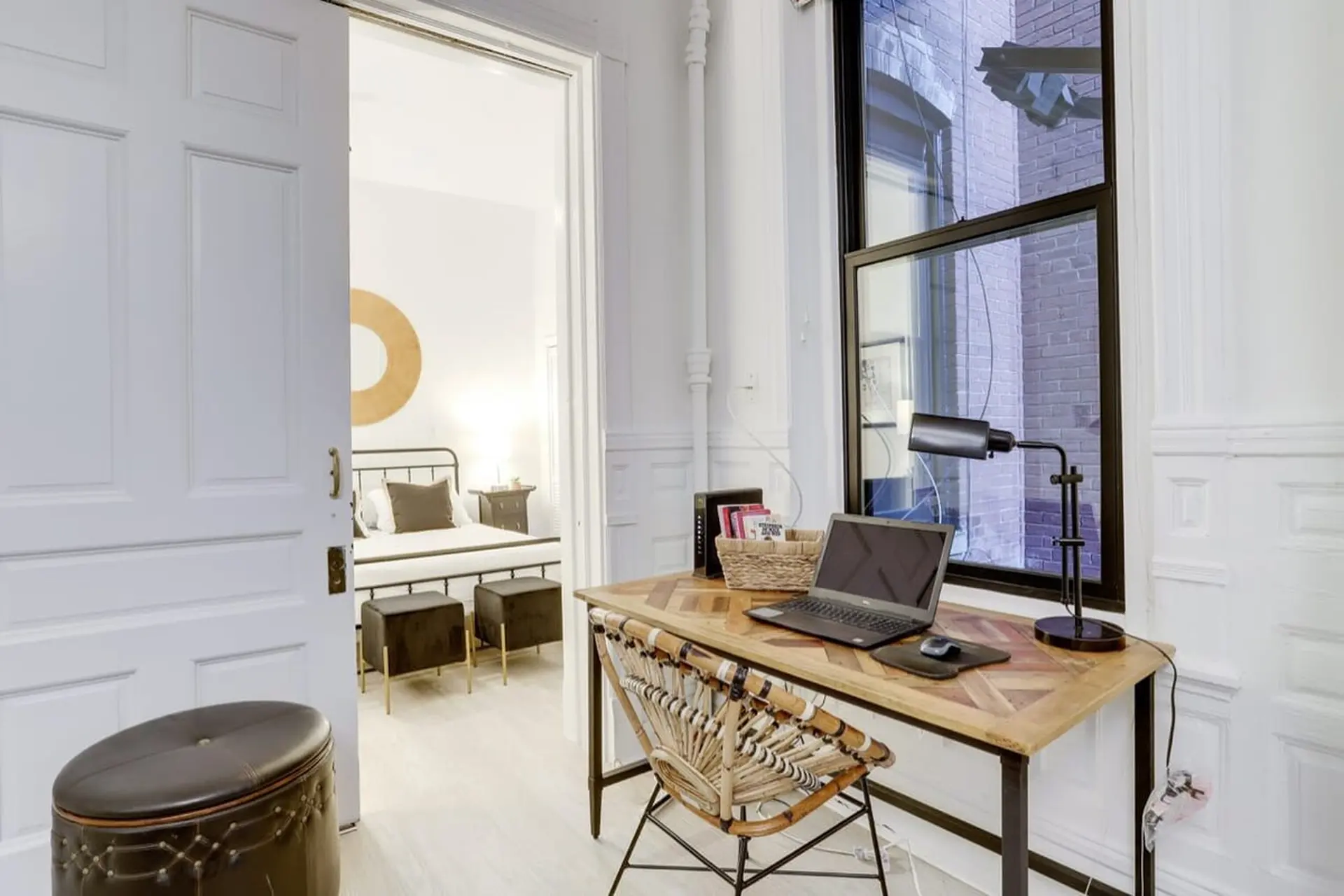 A desk with a laptop and lamp is in front of a window with a view of a brick building.
