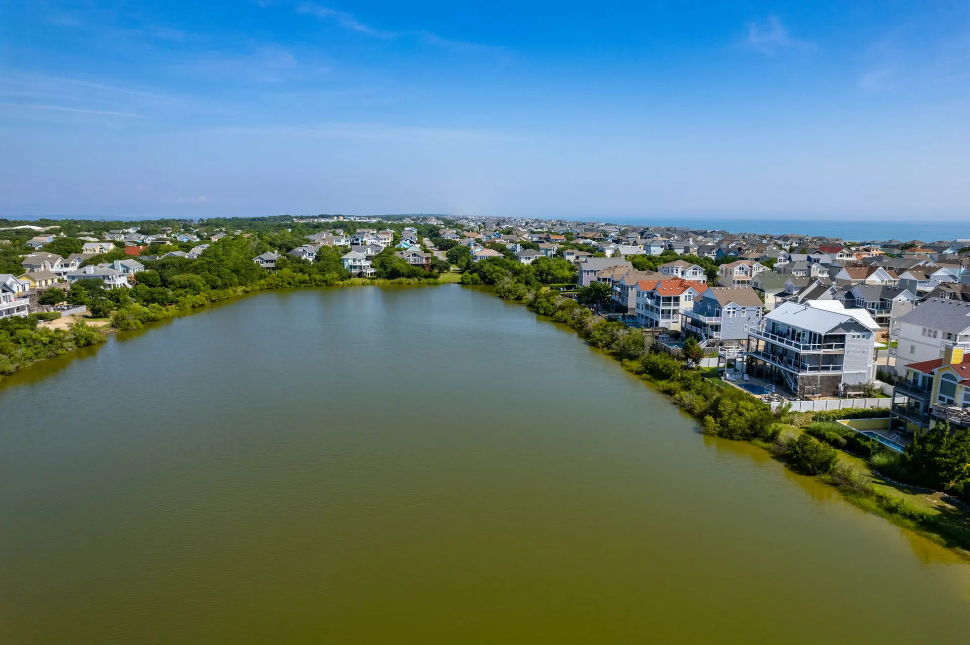 Aerial view of a lake surrounded by houses and trees, with the ocean in the background.