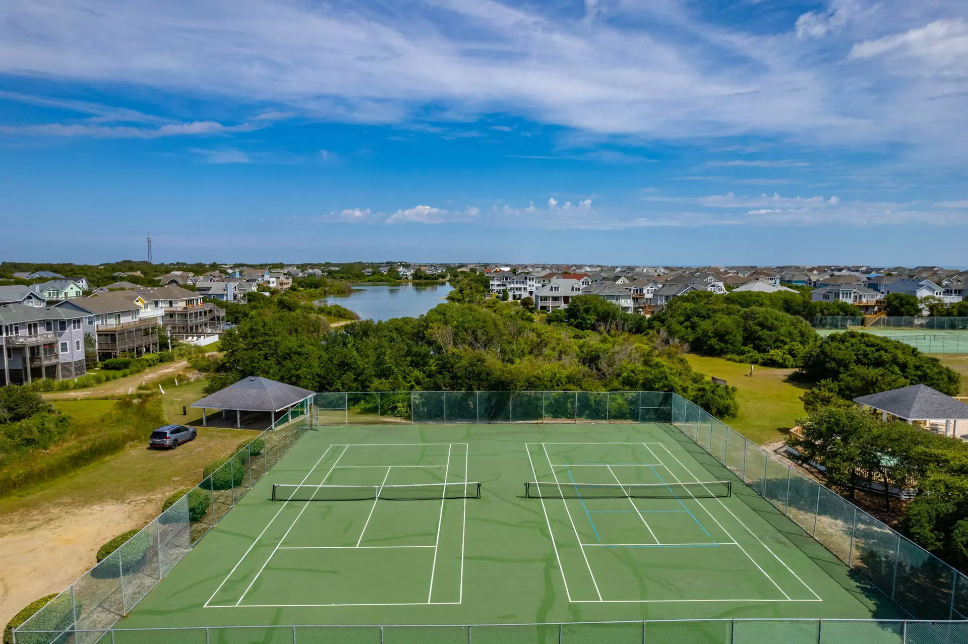 Tennis courts by a lake and houses under a blue sky with white clouds.