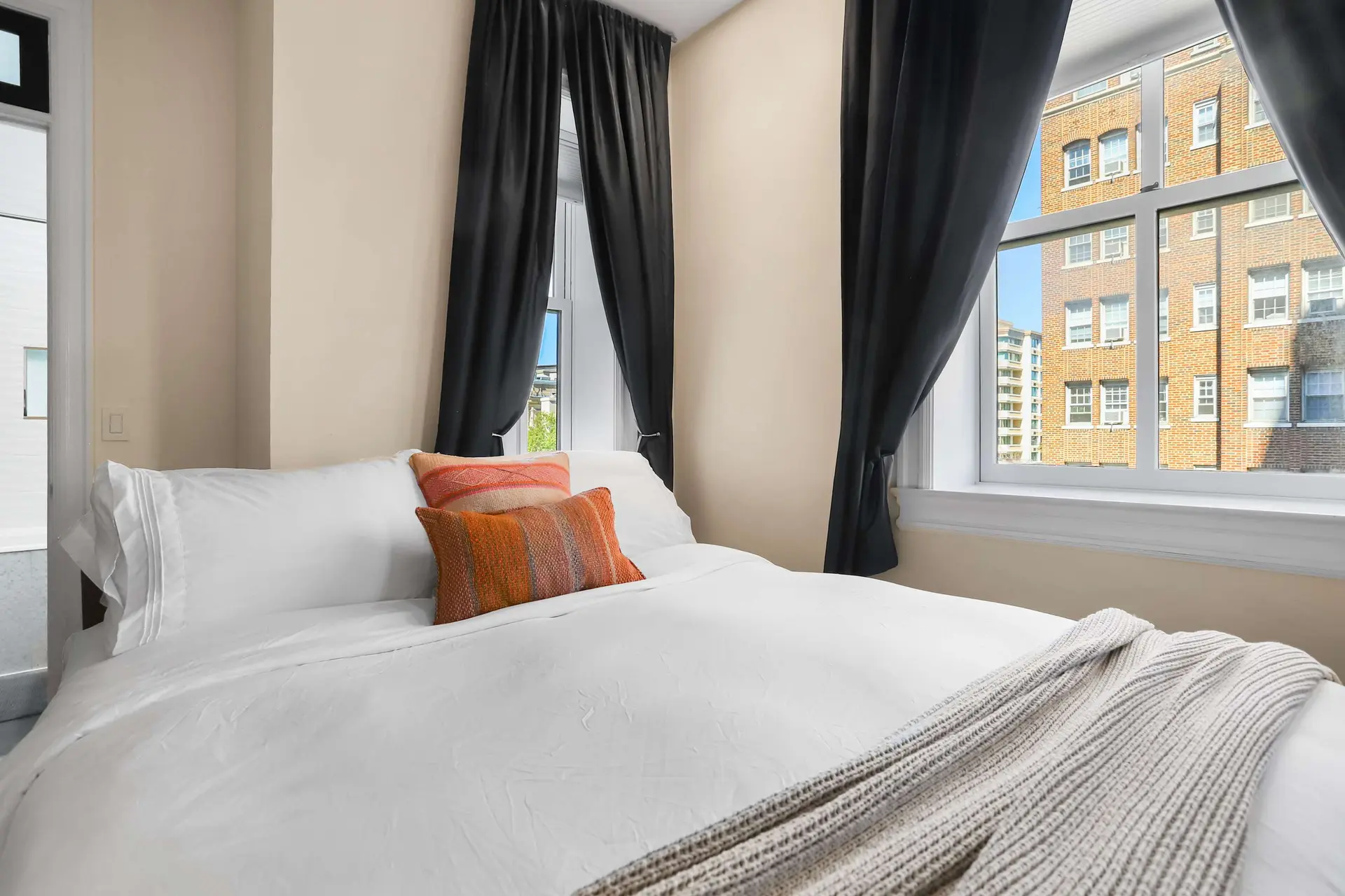 A bedroom with a white bed, dark curtains, and a window view of a brick building.