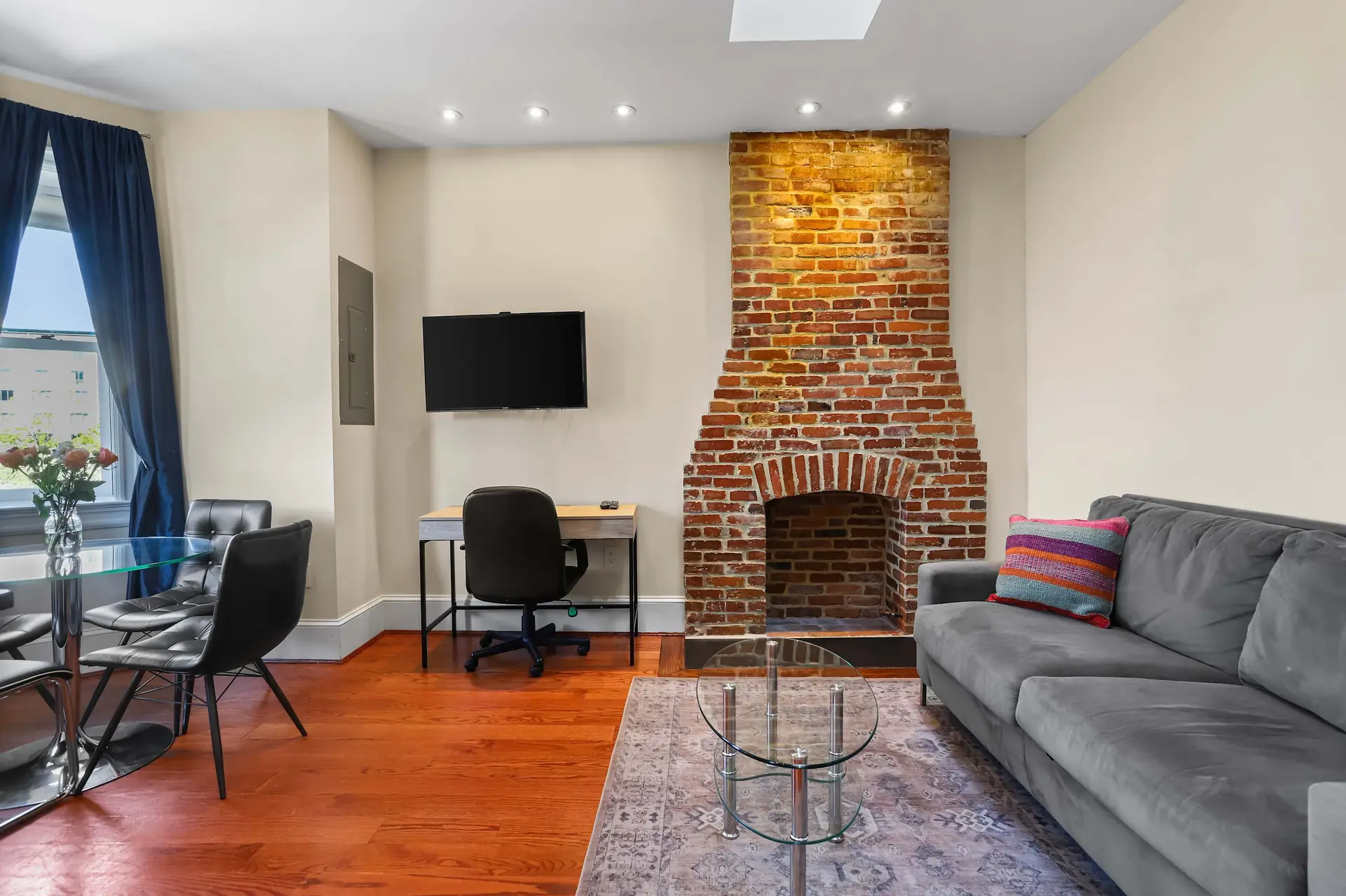 Living room with brick fireplace, gray sofa, glass coffee table, desk, and dining set.