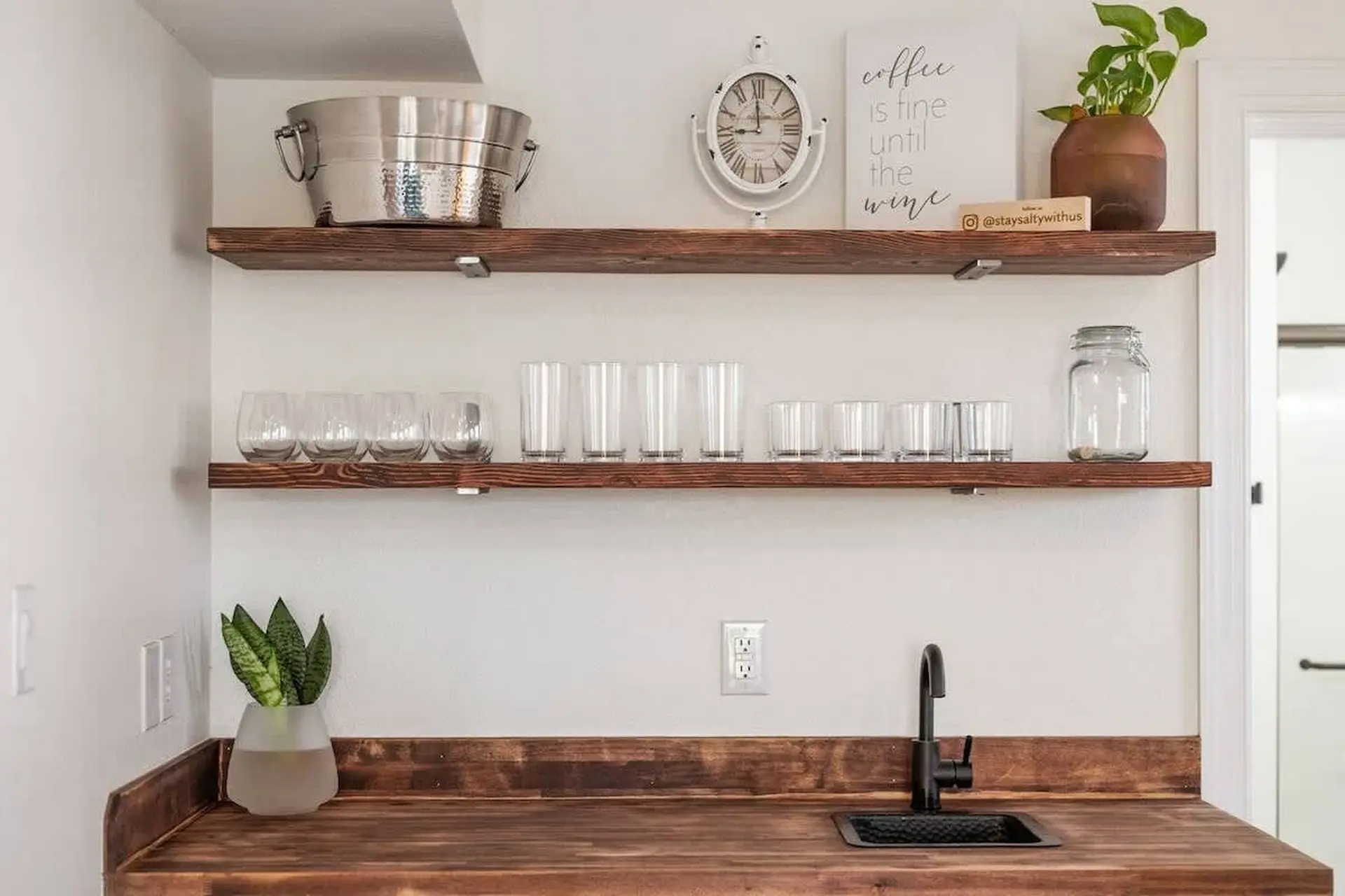 A wooden countertop with a sink and faucet, above it are two shelves with glasses, a bucket, a clock