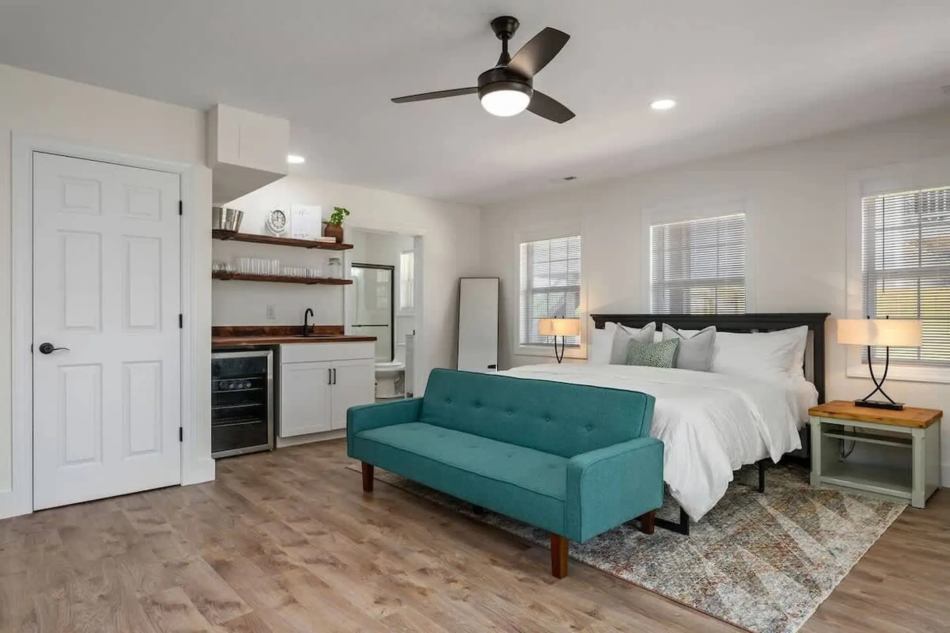 A modern bedroom with a teal sofa, king-size bed, and a small kitchenette.