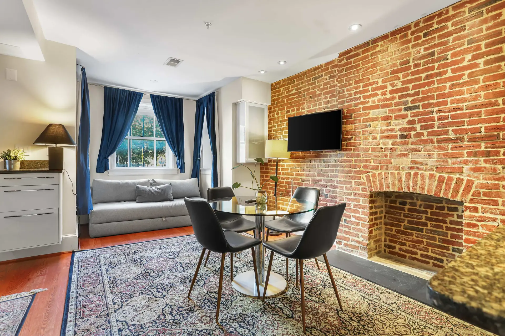 A round glass dining table with black chairs is in front of a brick wall with a fireplace.