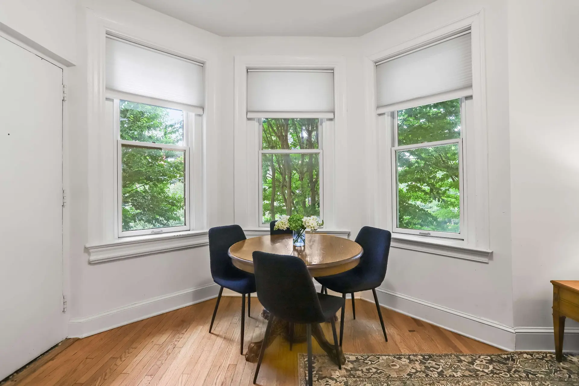 A round dining table with four chairs is set in a bay window.
