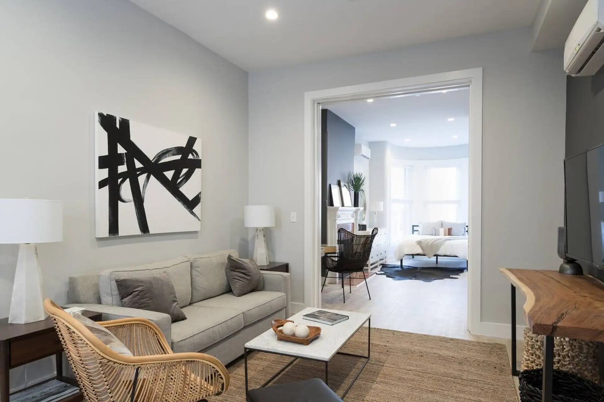 Living room with neutral tones, modern art, and woven furniture, opening into a bedroom.