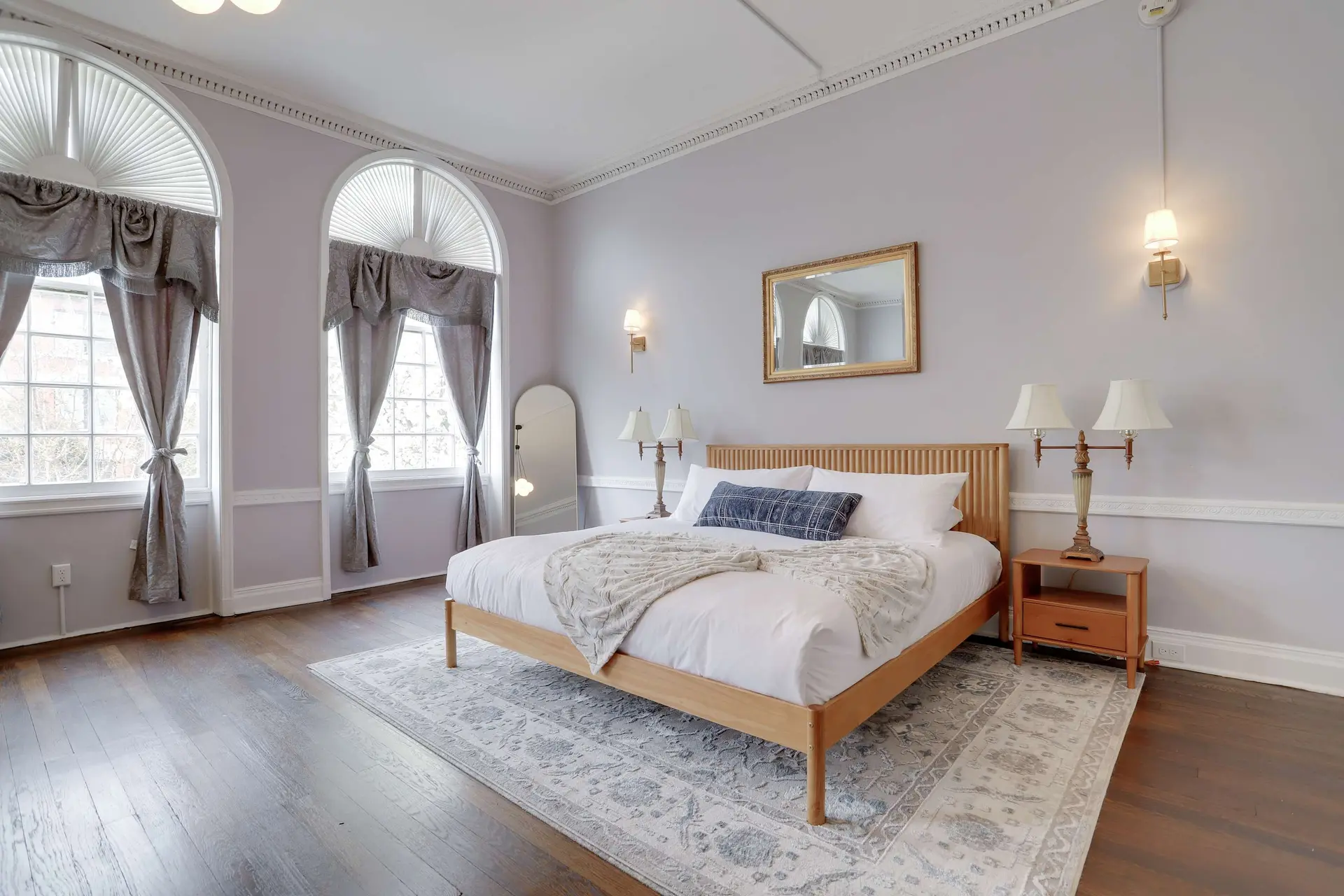 A bedroom with a large bed, two arched windows with ornate curtains, and a golden mirror.