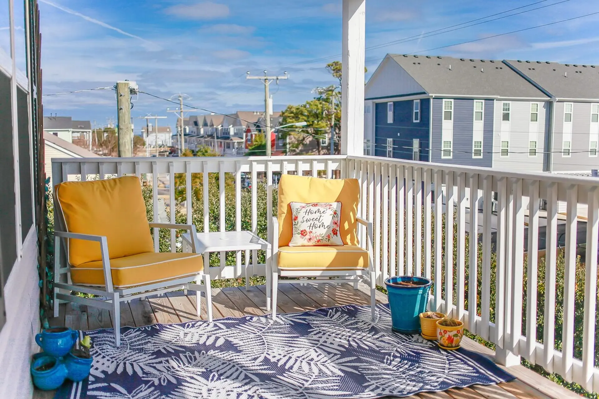 A porch with two yellow chairs, a rug, and potted plants overlooks buildings and a street.