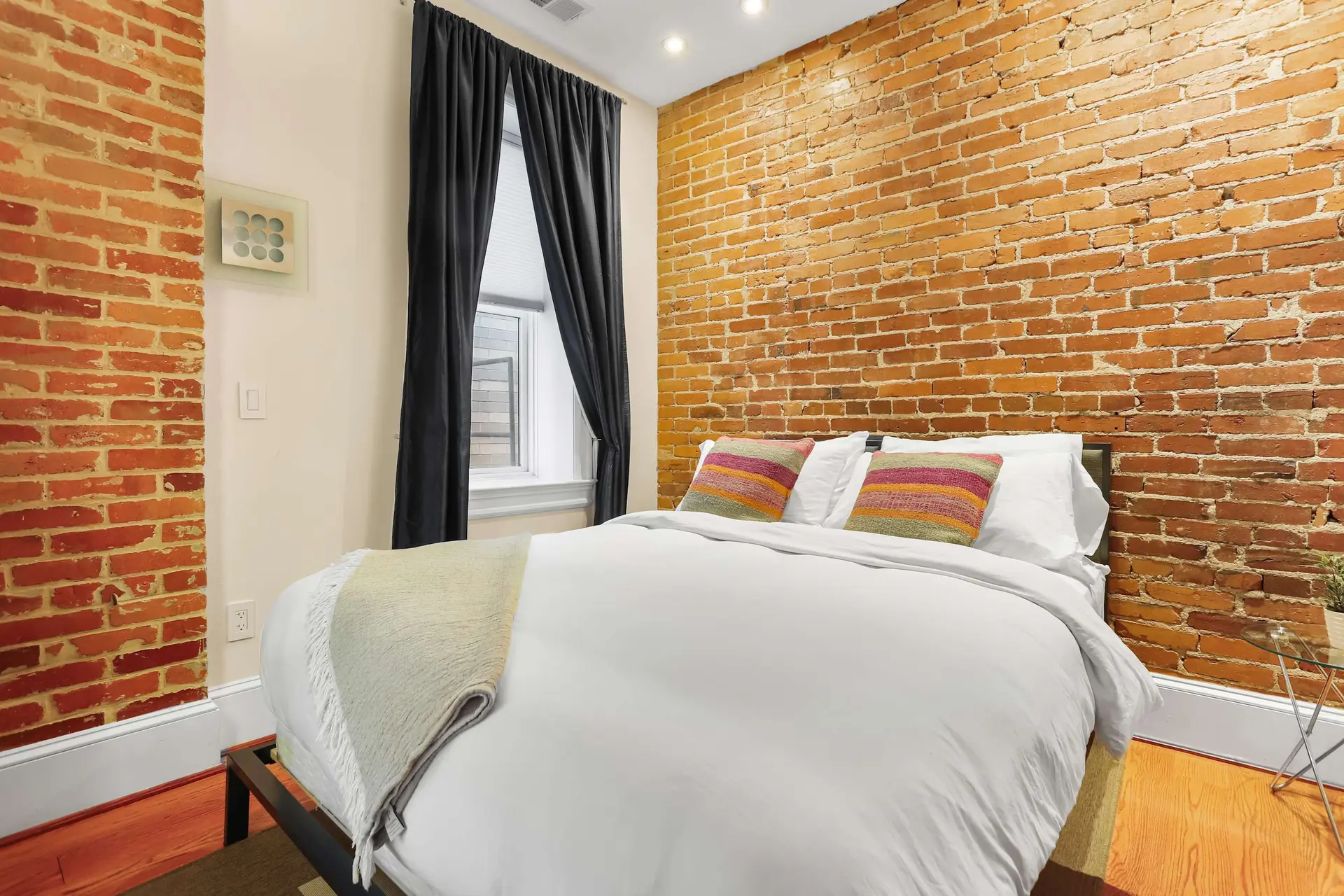 A cozy bedroom with exposed brick walls, a white duvet, and colorful pillows.
