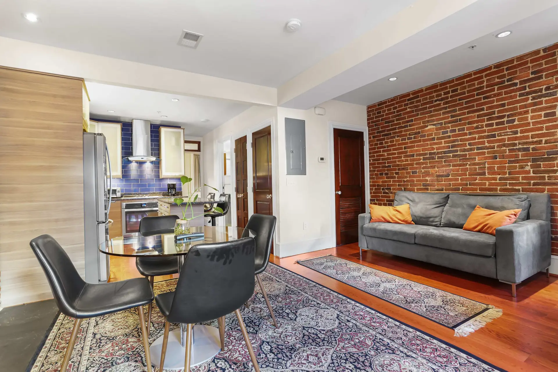 A gray couch sits in front of a brick wall, with a glass dining table and chairs nearby.