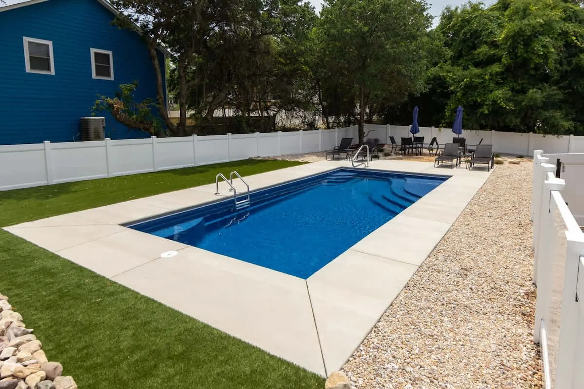 A bright blue swimming pool with steps and a ladder is surrounded by green artificial turf, concrete