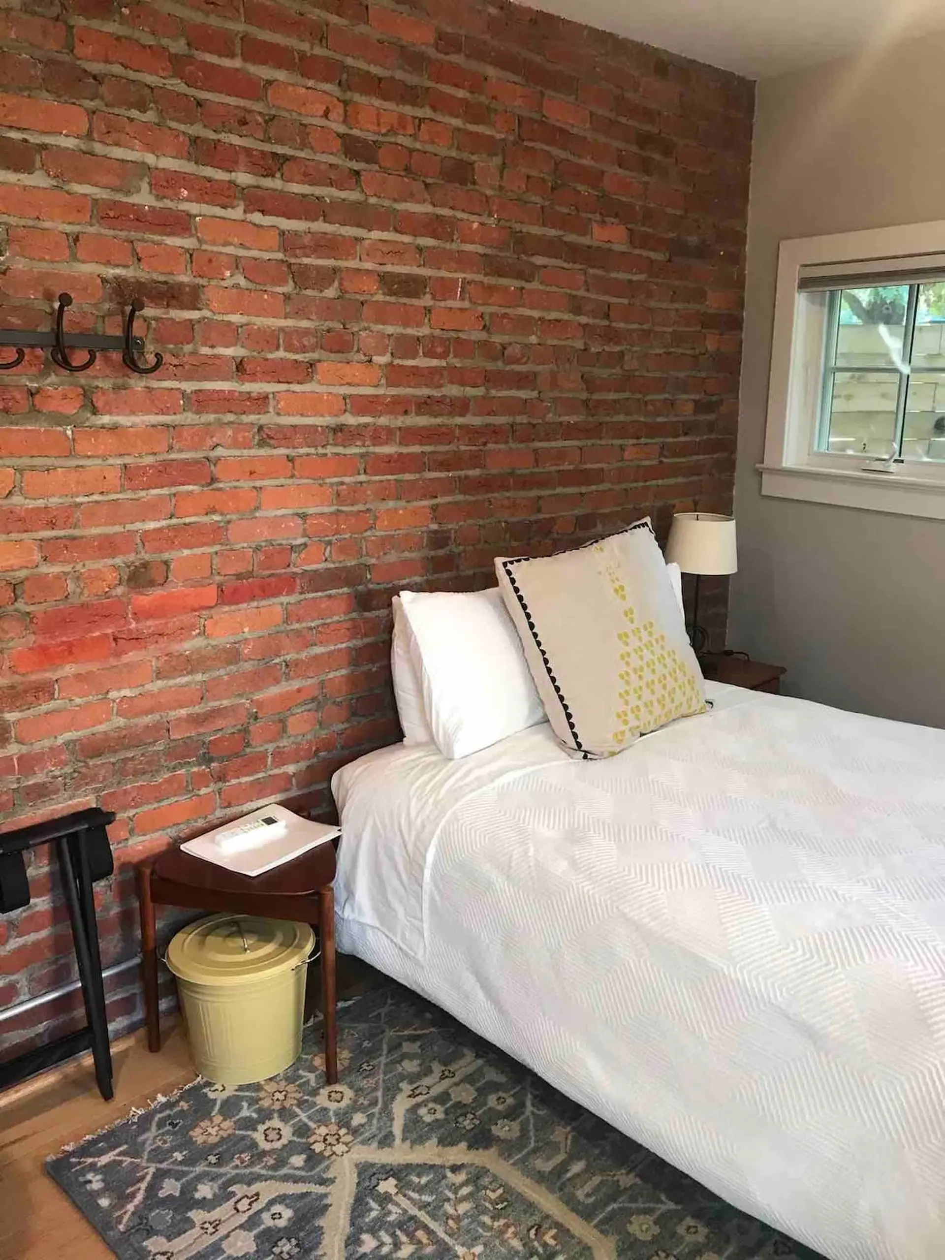 A bed with white bedding and a decorative pillow sits next to a brick wall, a nightstand, and a wind