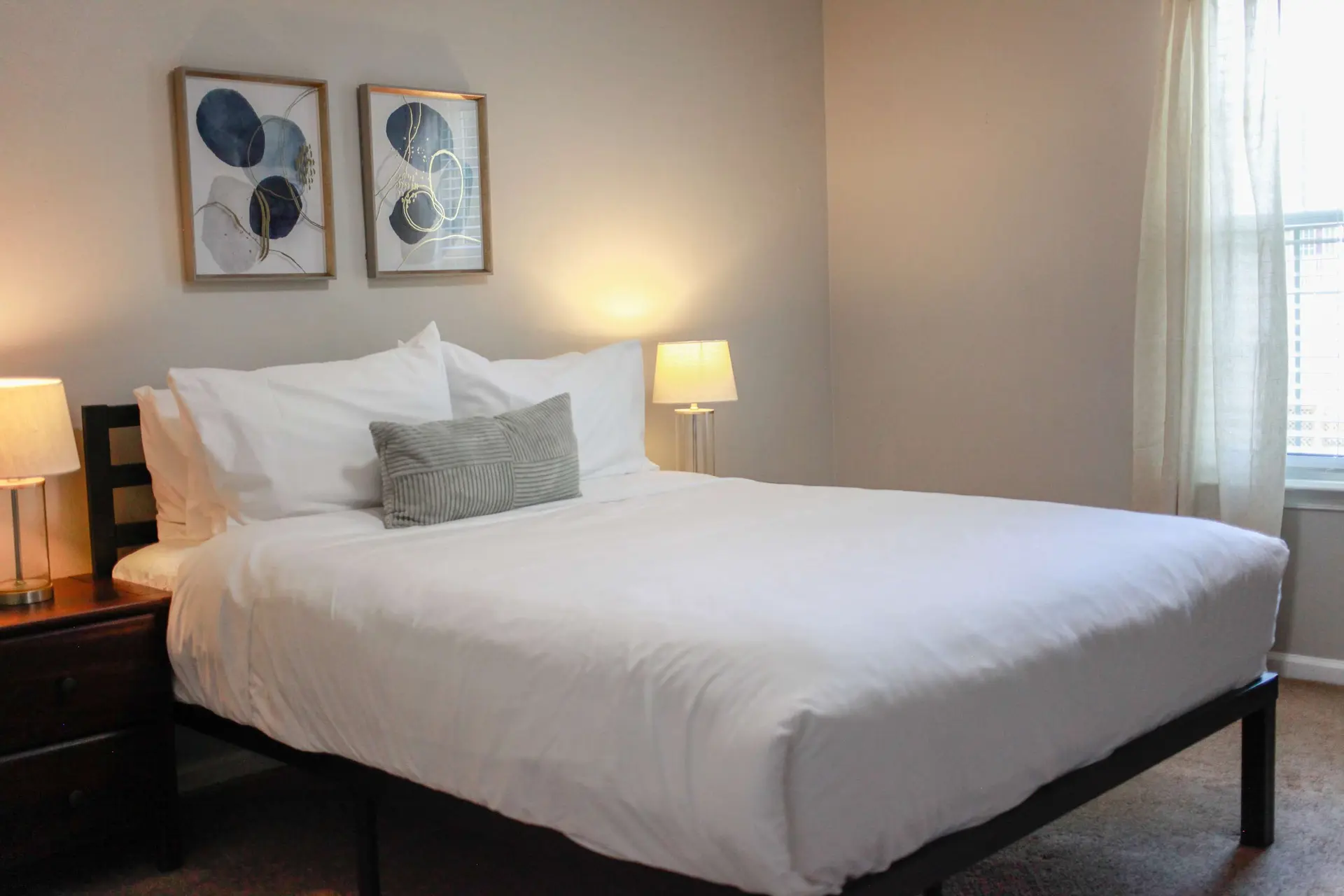 A white bed with two pillows, a grey pillow, and two framed art pieces hangs on the wall.