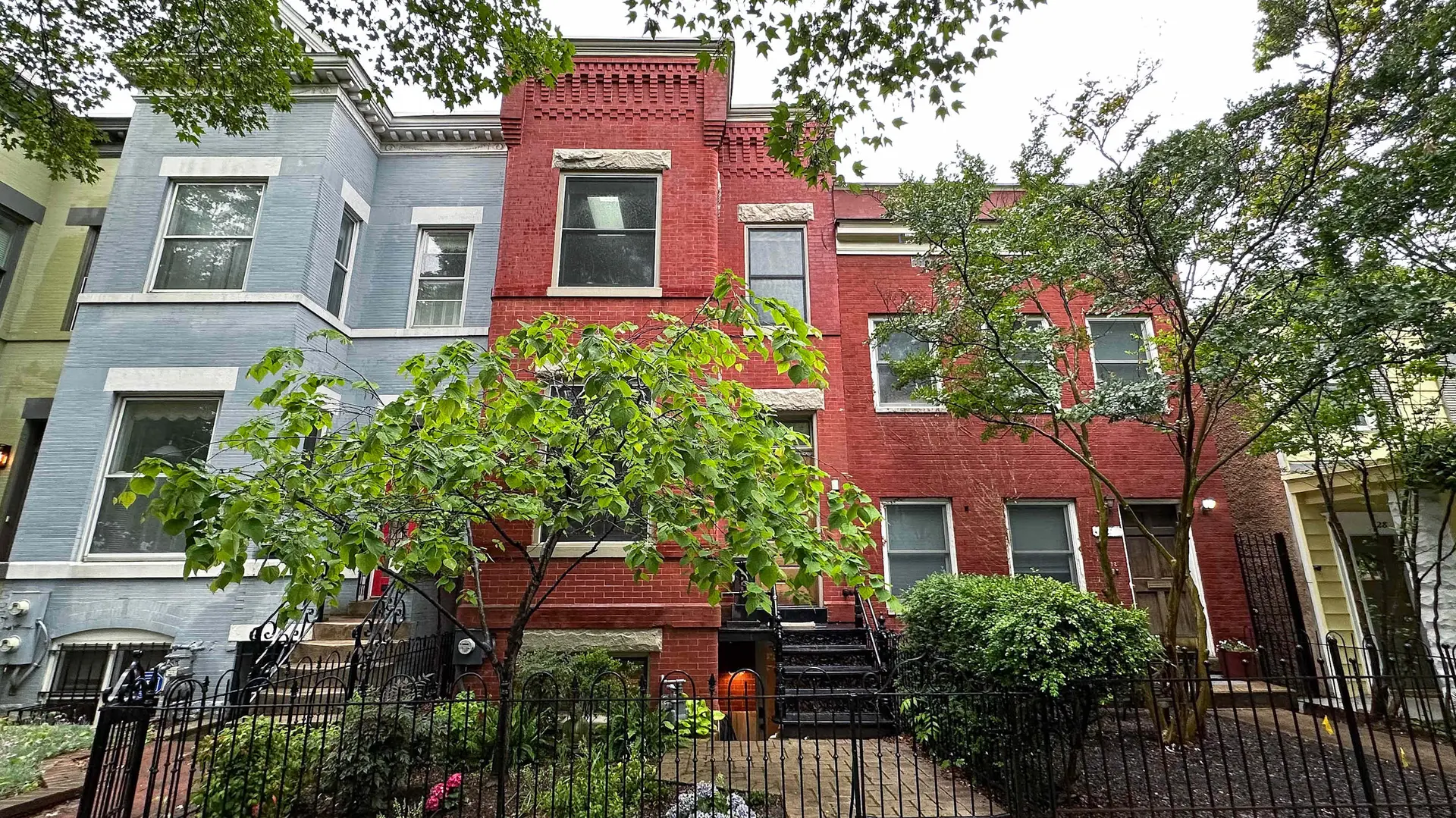 A light blue rowhouse sits next to a red brick rowhouse with a small tree in front.