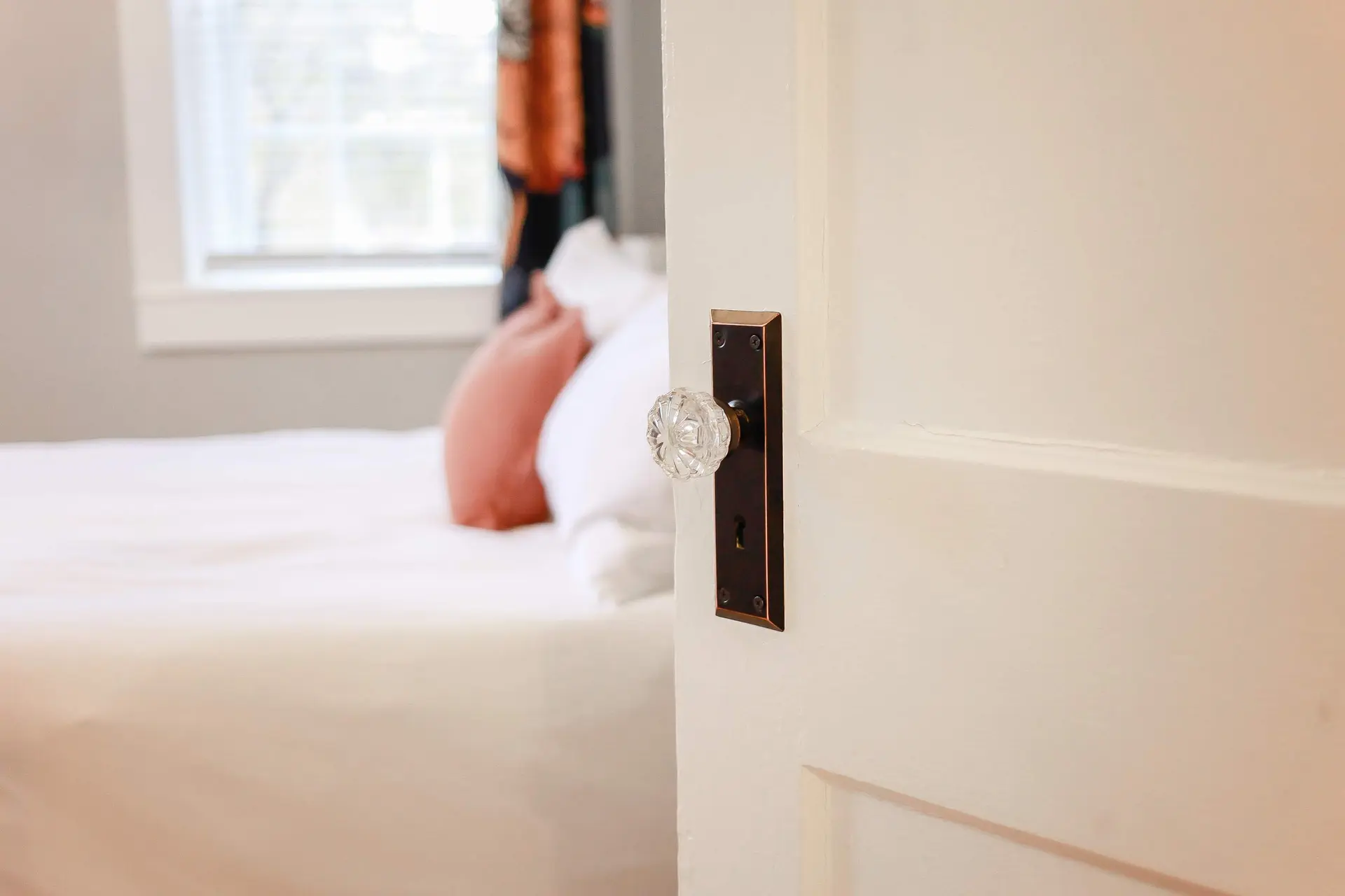 A slightly open door with a crystal doorknob reveals a blurred bed with white and coral pillows.