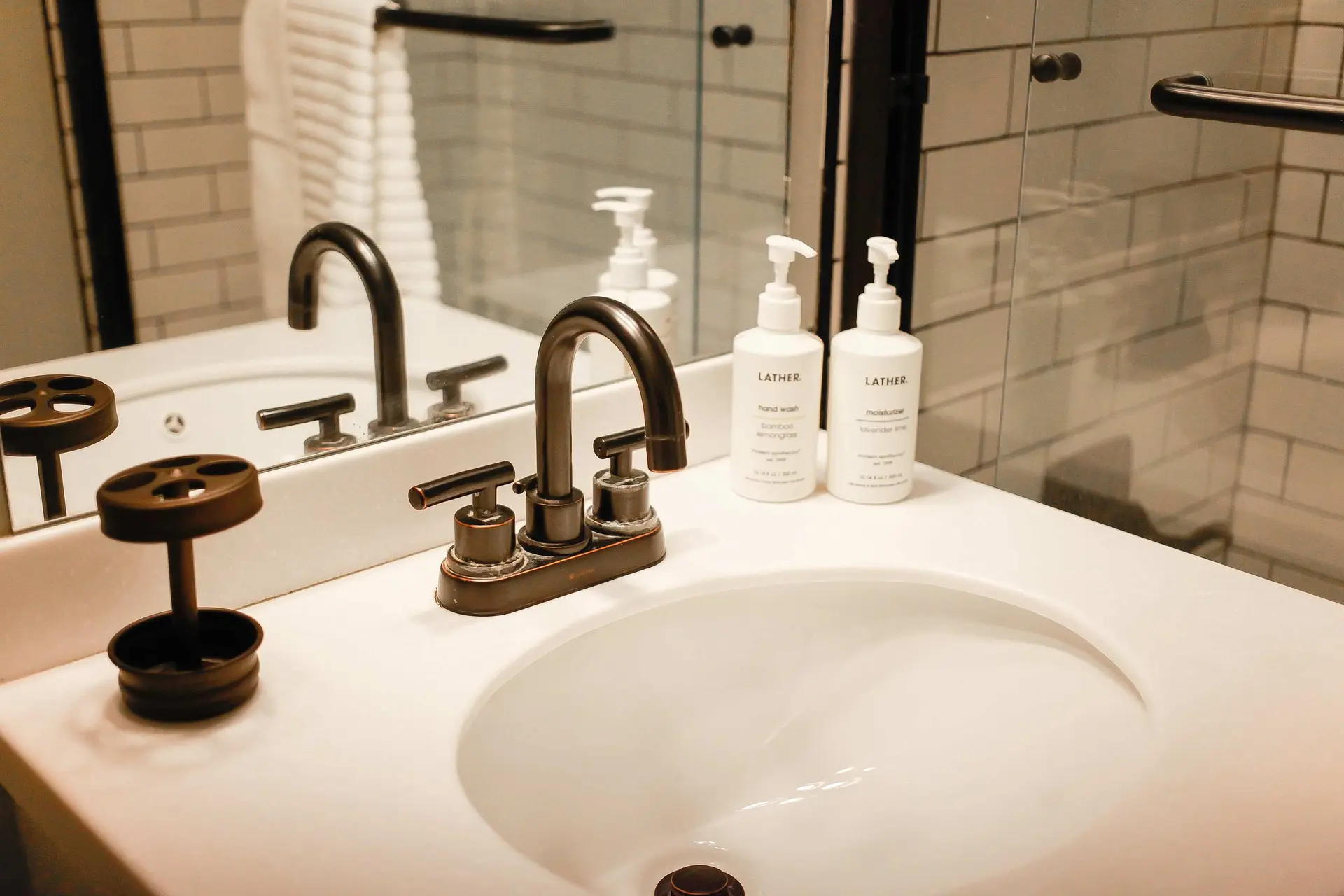 A sink with two soap dispensers and a faucet, with a tiled wall and mirror in the background.